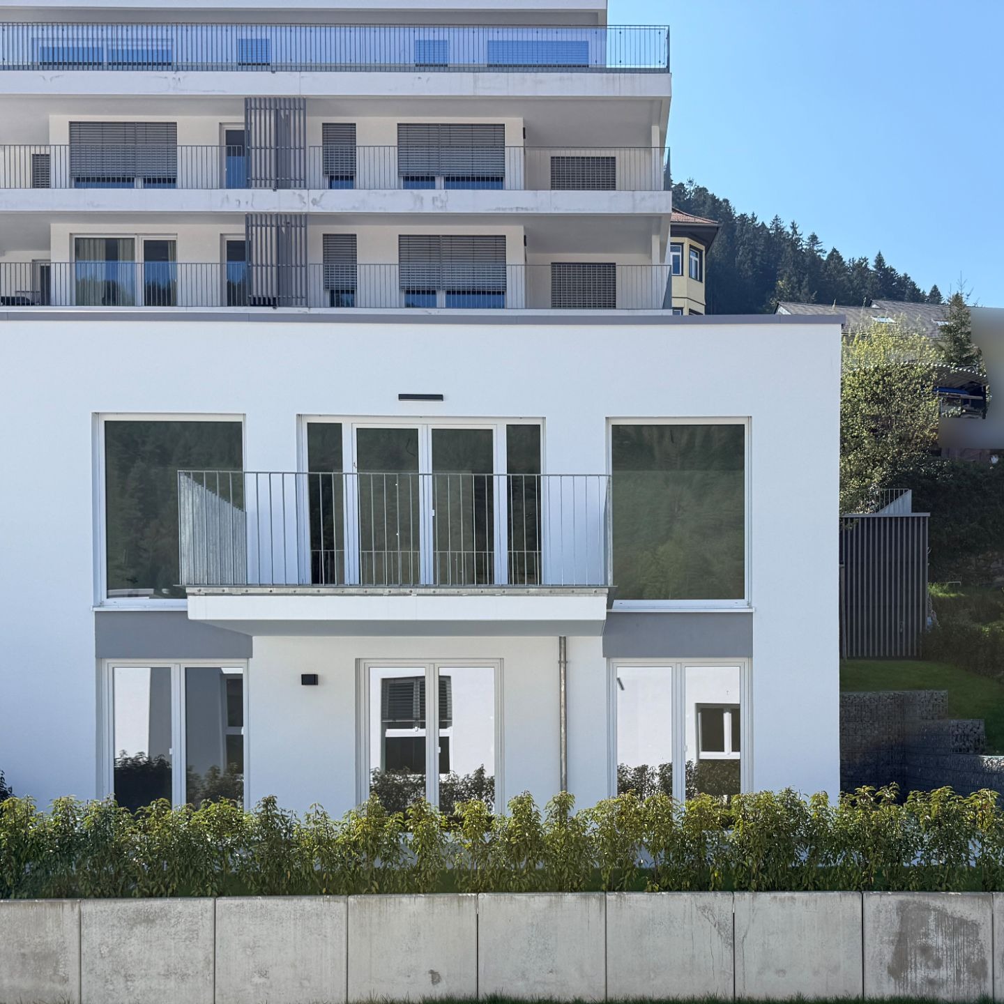 Modernes weißes Wohnhaus mit großen Fenstern und Balkon vor einer Hecke.