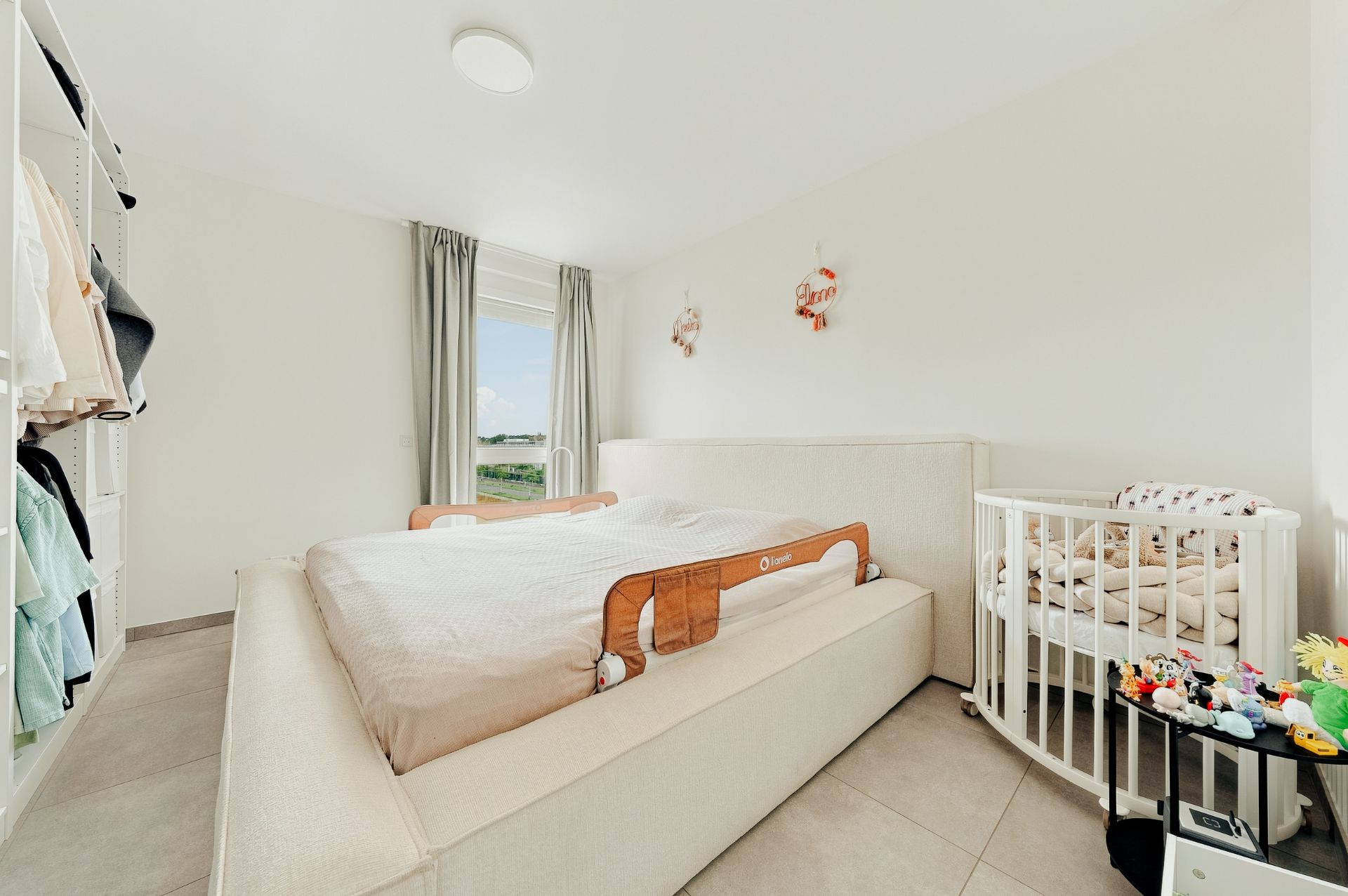 Schlafzimmer mit großem Bett, Babybett und offenen Kleiderschränken, hell und minimalistisch eingerichtet.