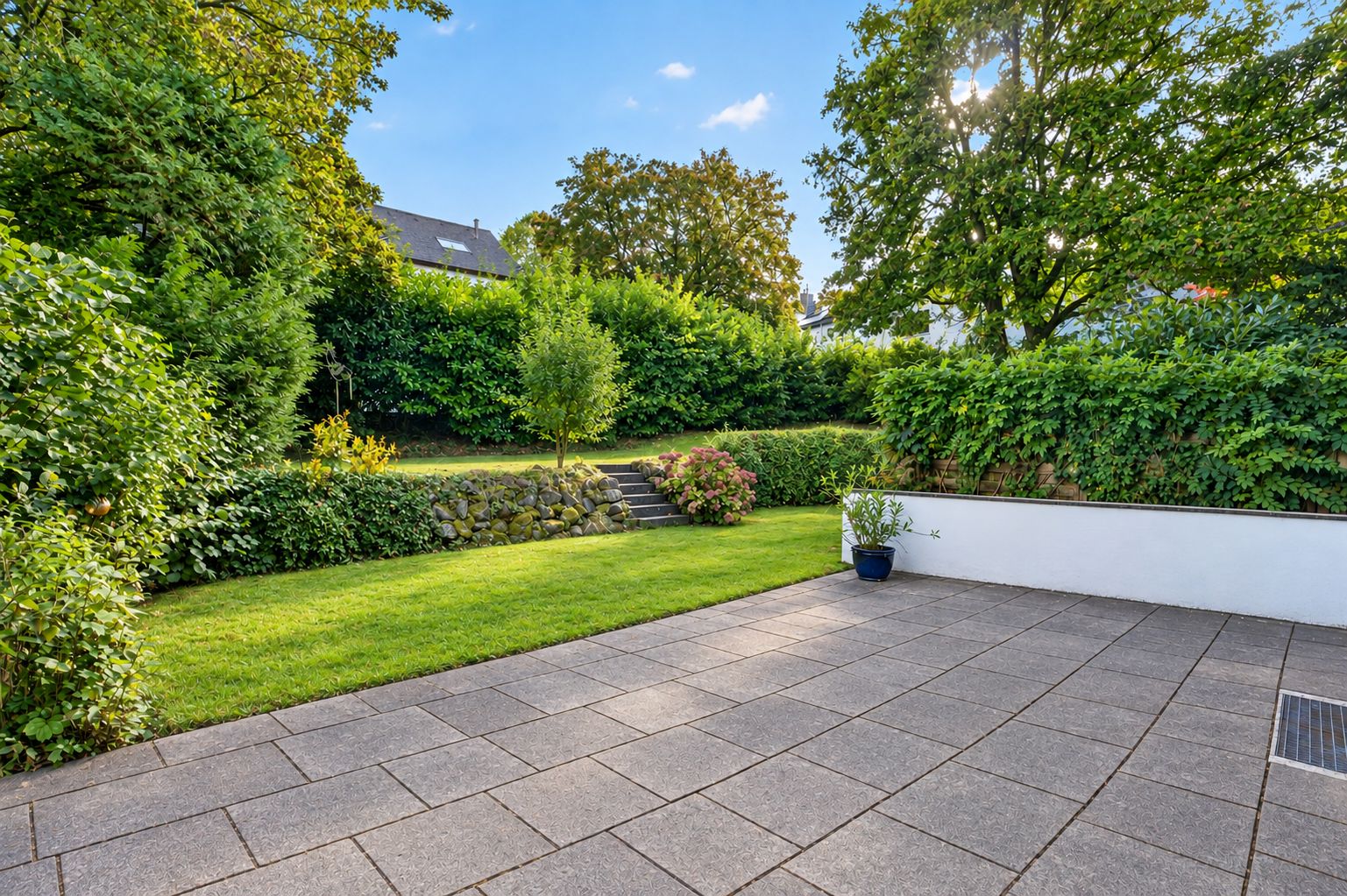 Große Terrasse mit Fliesenboden, angrenzender Rasenfläche und Bäumen im Hintergrund.