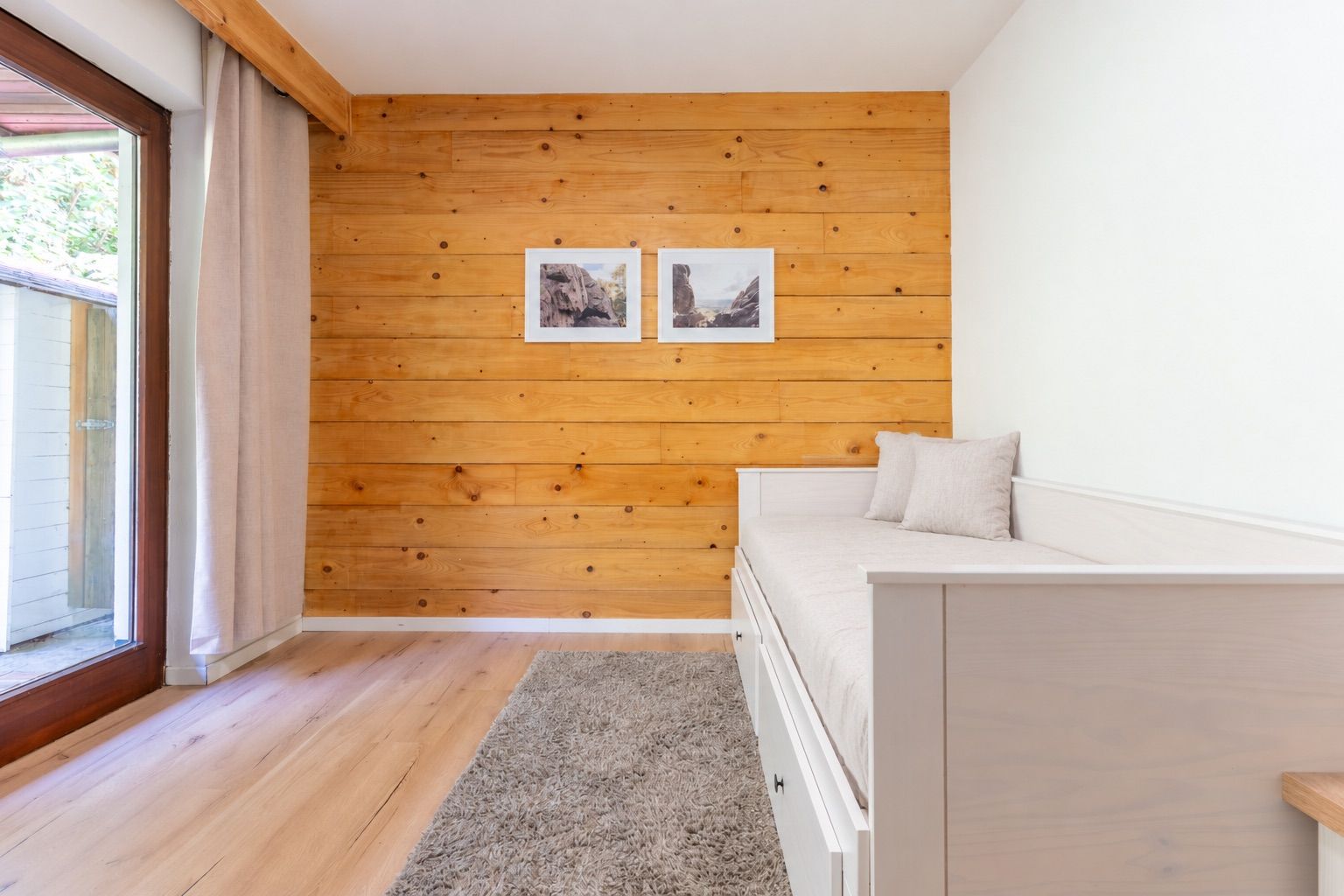 Helles Zimmer mit Holzwand, Tagesbett, Teppich und großem Fenster mit Vorhang.