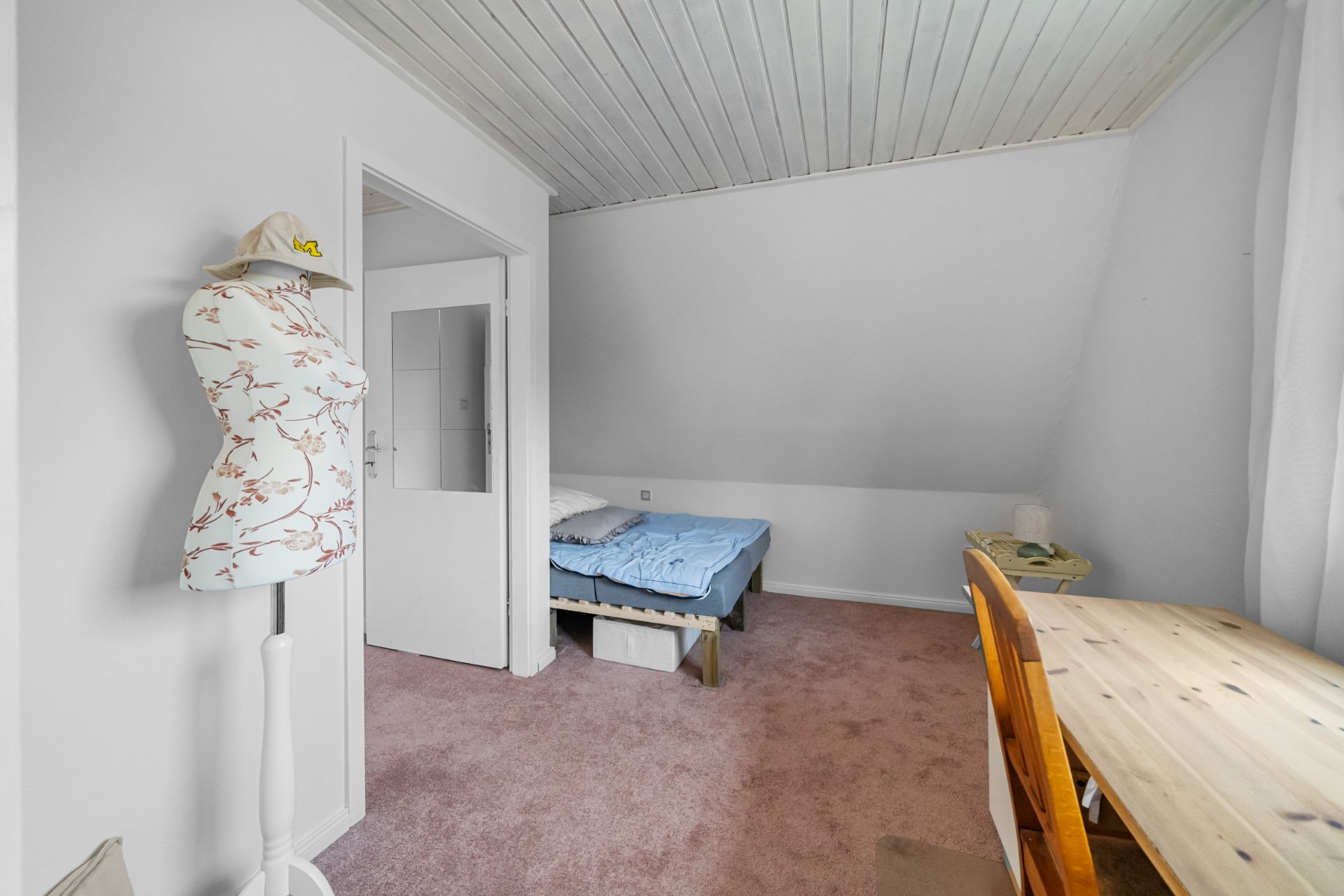 Zimmer mit Schreibtisch, Stuhl, Schneiderpuppe und Bett unter einer Dachschräge.