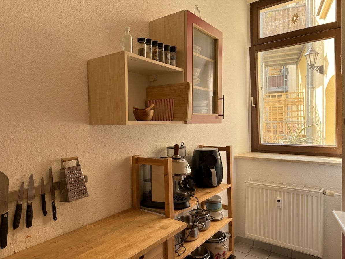Kleine Küche mit Holzregalen, Messern, Kaffeemaschine und Fenster bei Tageslicht.