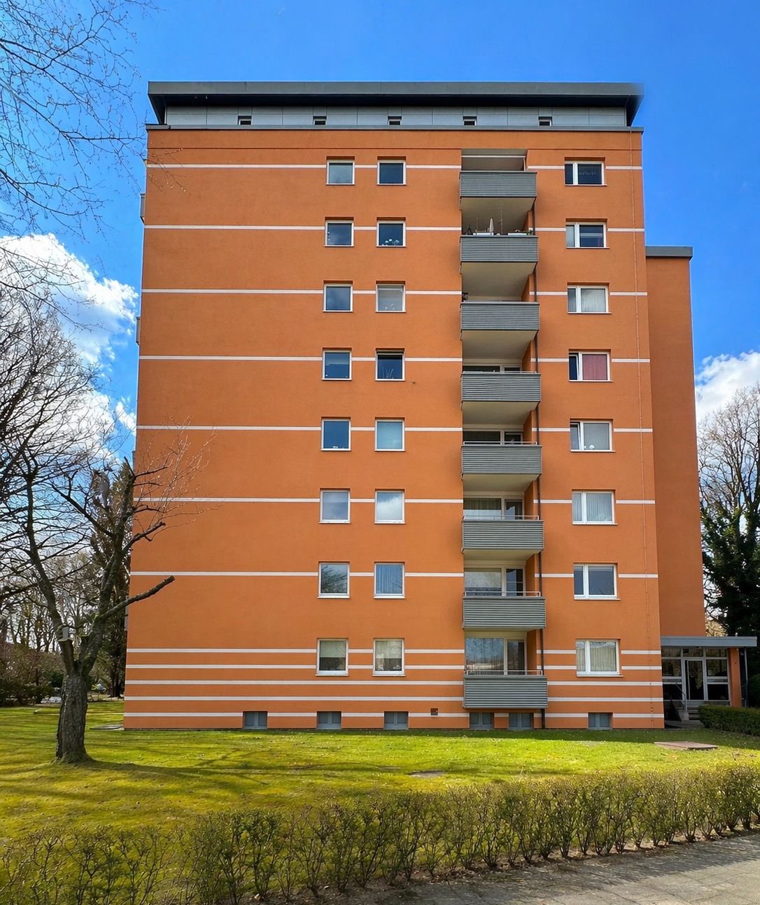 Orangefarbenes Wohnhaus mit mehreren Balkonen und Fenstern, davor Rasen und Bäume.