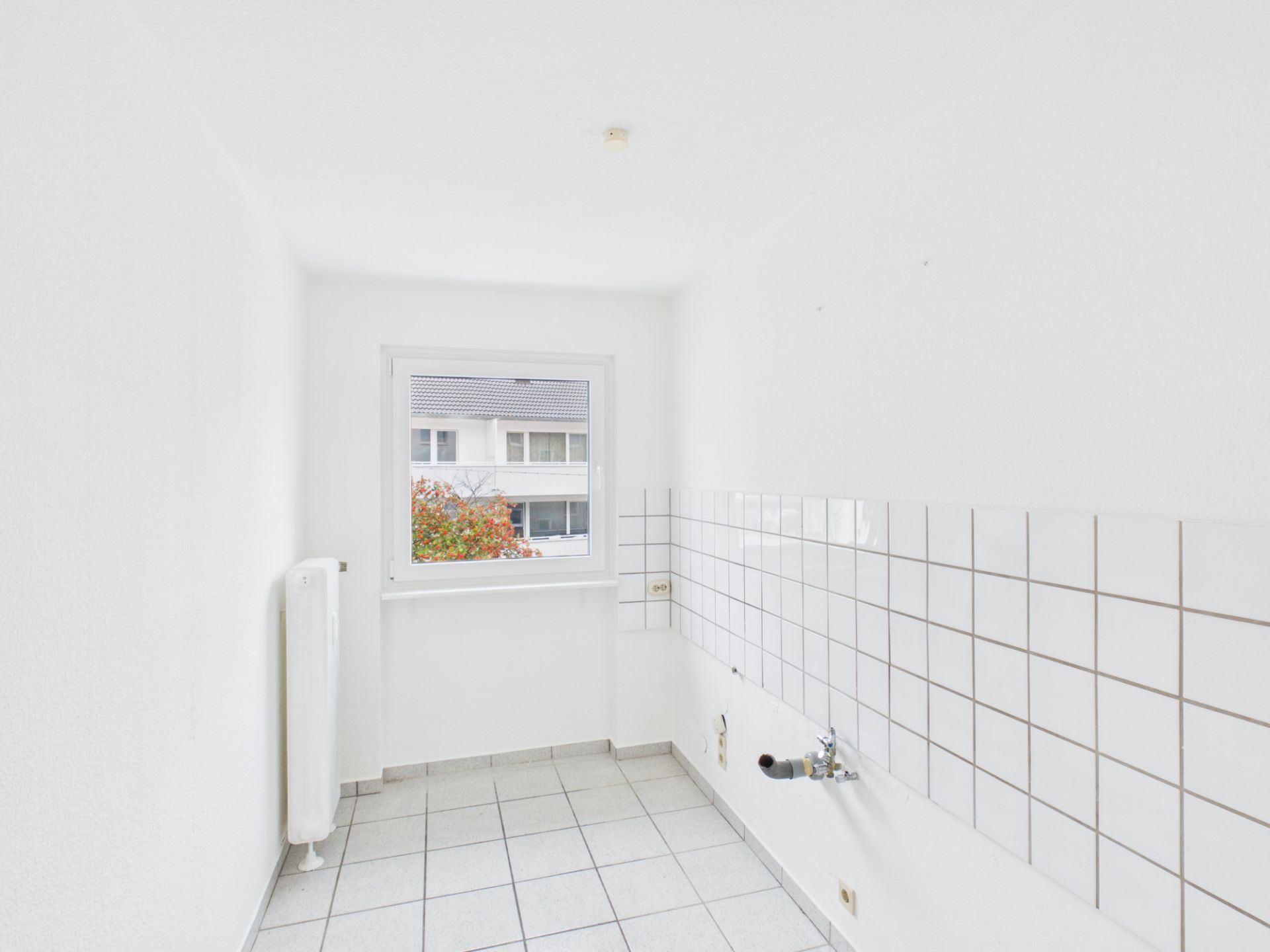 Heller, leerer Raum mit Fliesenboden, Fenster und weißen Wandfliesen, Heizung links.
