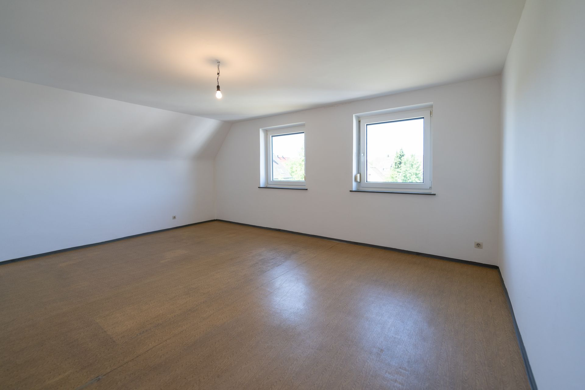 Leeres Zimmer mit zwei Fenstern, Holzboden und weiß gestrichenen Wänden.