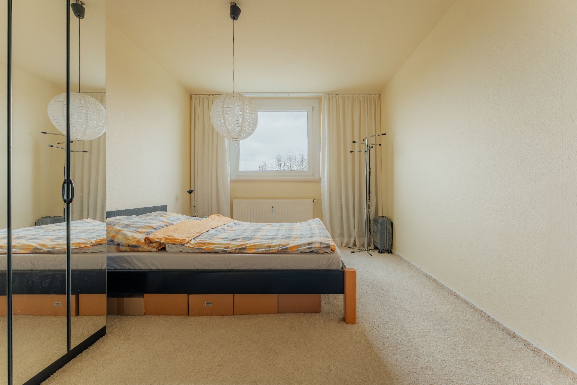 Schlafzimmer mit Doppelbett, Spiegelkleiderschrank und Fenster mit weißen Vorhängen.
