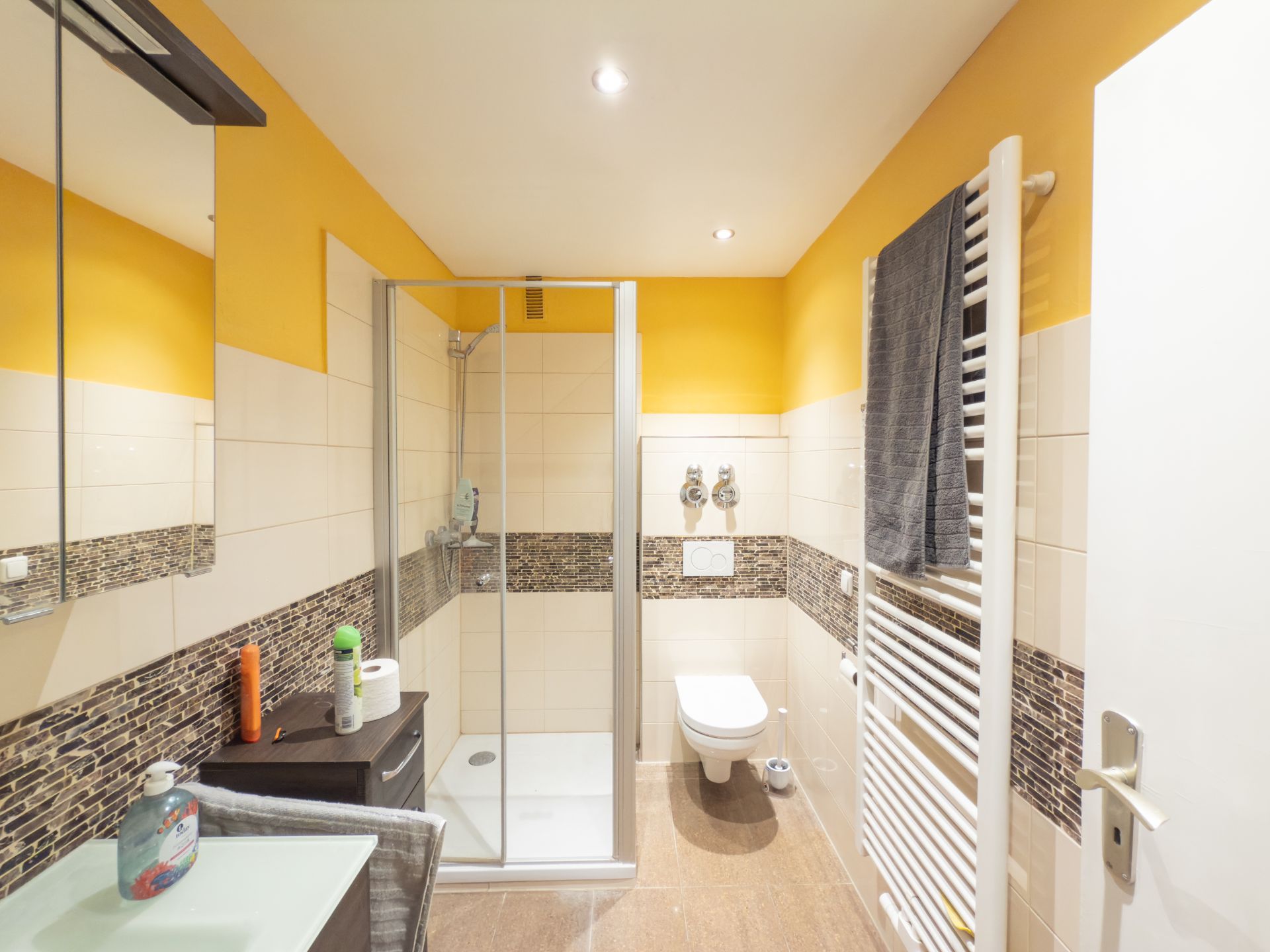 Modernes Badezimmer mit Dusche, WC, Waschbecken und Handtuchheizkörper, Wände teils gelb gestrichen.