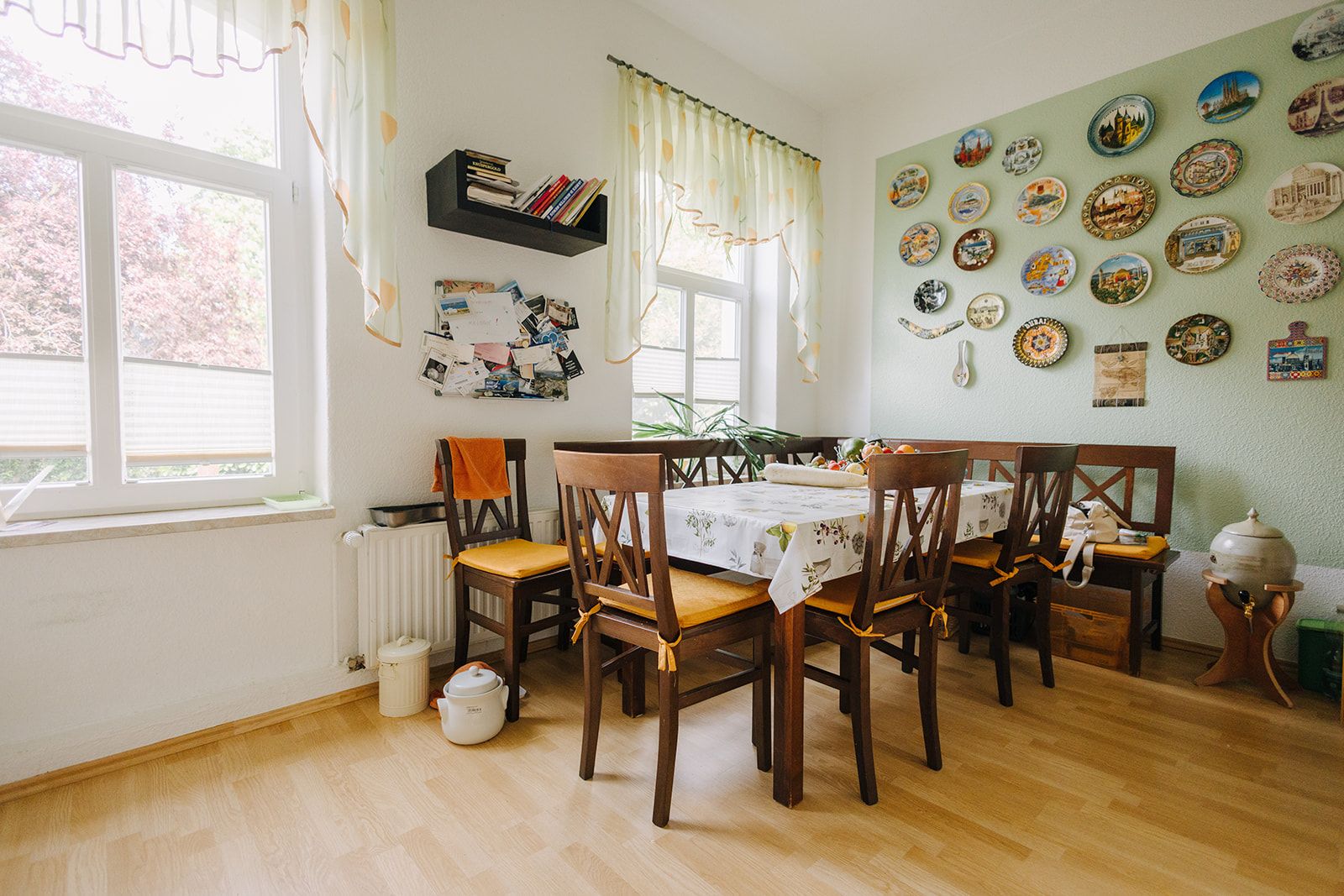 Esszimmer mit Holztisch, sechs Stühlen, Wandtellern und großen Fenstern mit Vorhängen.