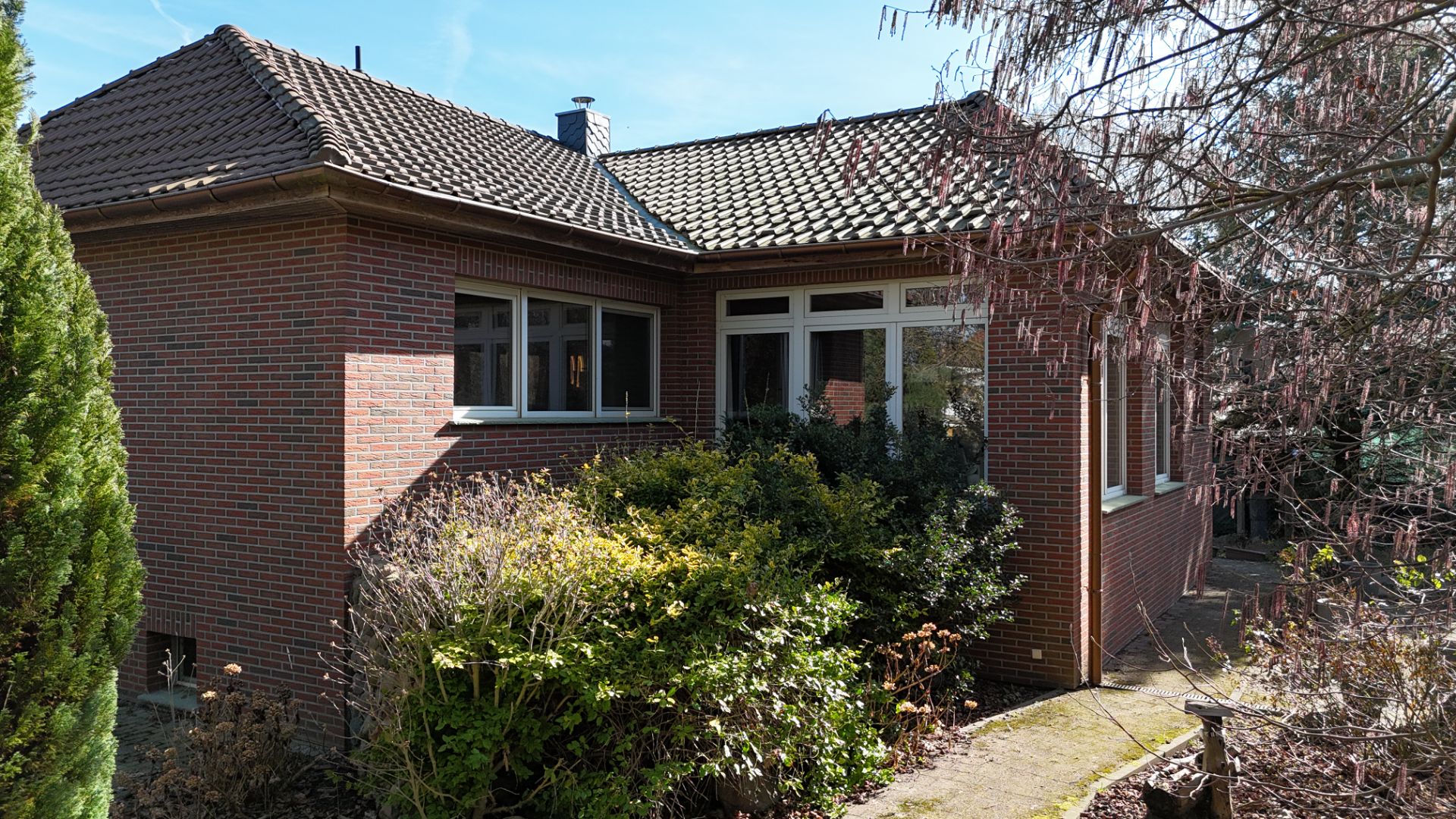 Backstein-Einfamilienhaus mit Satteldach und Garten, vorne Sträucher und ein schmaler Weg.