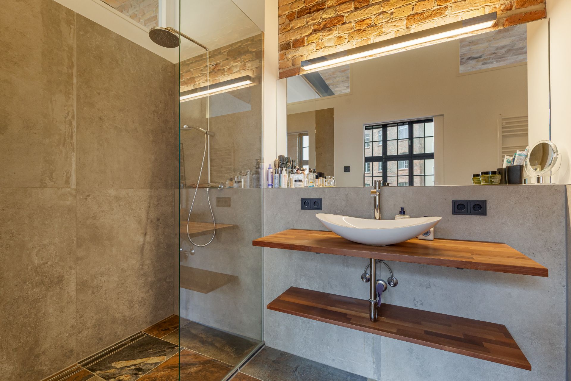 Modernes Bad mit Glasdusche, Waschbecken auf Holzplatte und Spiegel vor Backsteinwand.