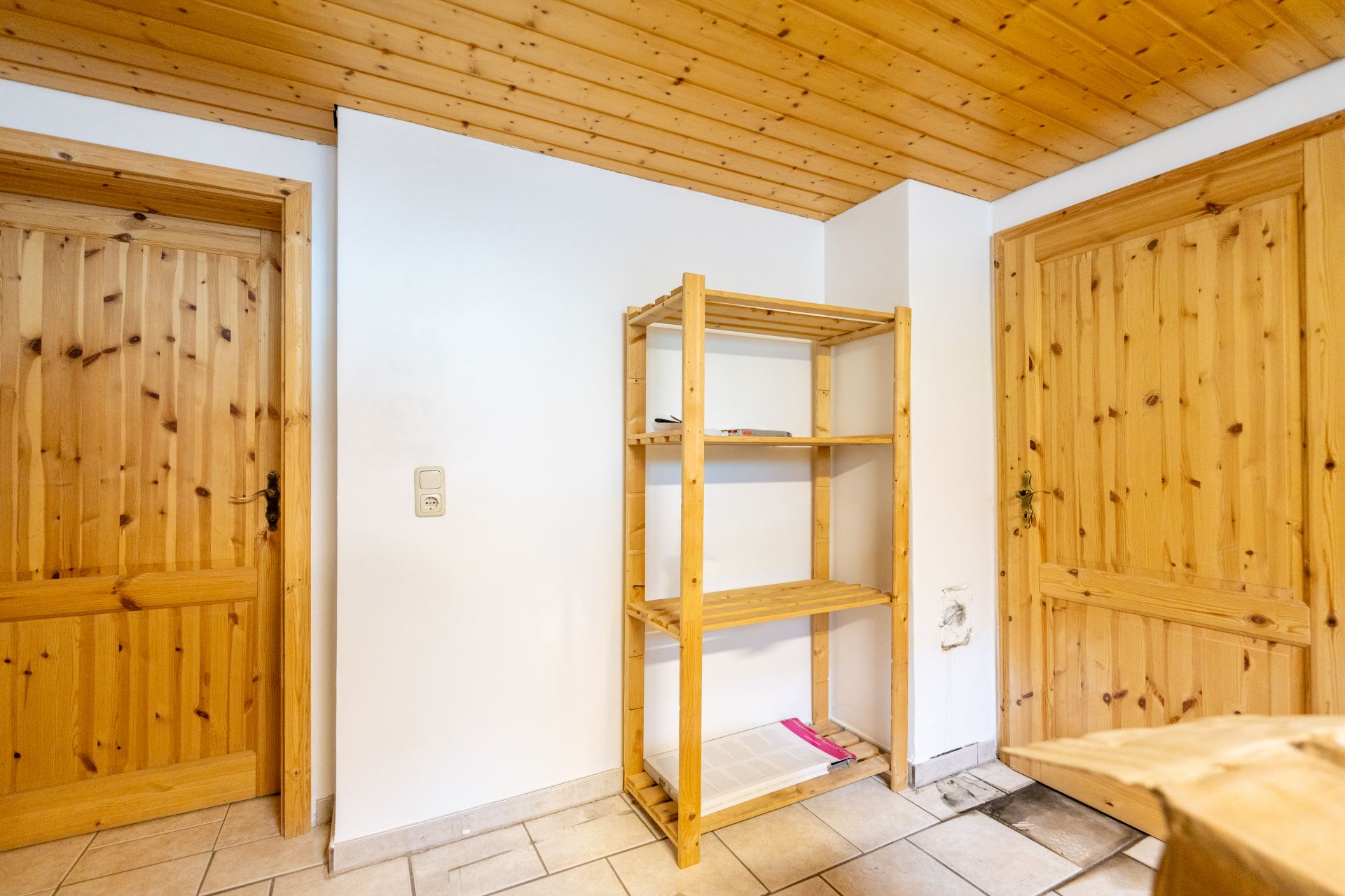 Raum mit Holztüren, Holzregal und gefliesten Boden, helle Wände und Holzdecke.