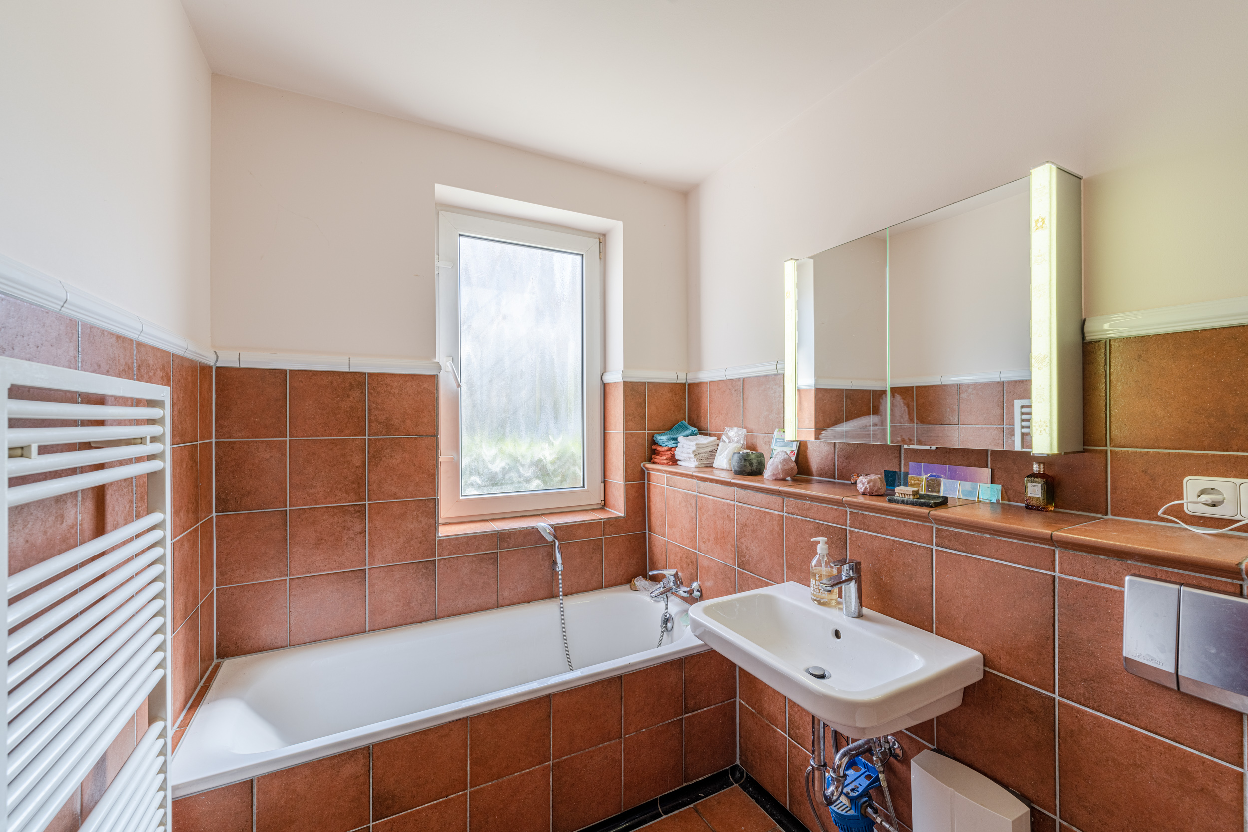 Badezimmer mit braunen Fliesen, Badewanne, Waschbecken, Fenster und beleuchtetem Spiegel.