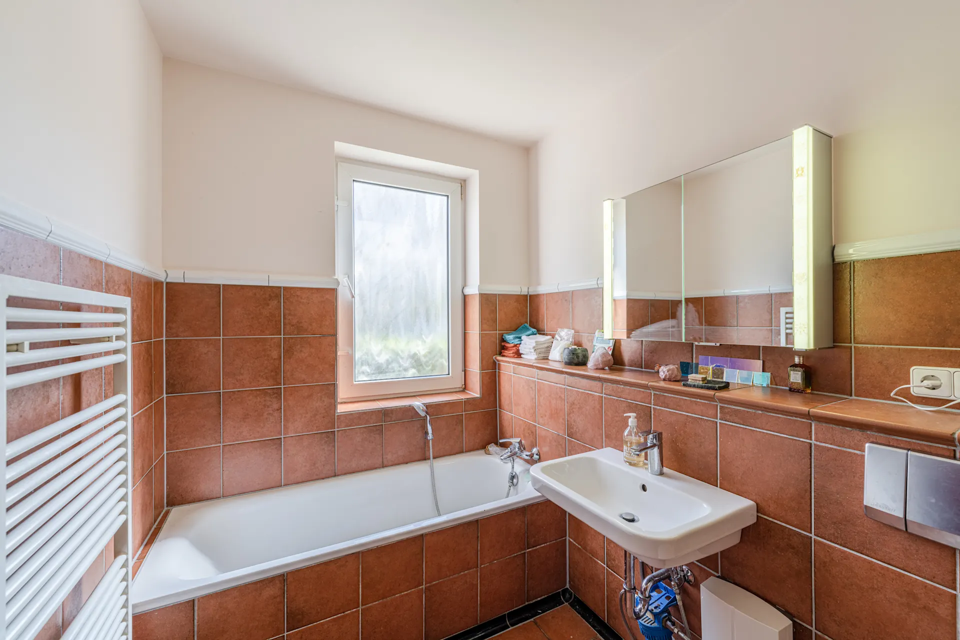 Badezimmer mit braunen Fliesen, Badewanne, Waschbecken, Fenster und beleuchtetem Spiegel.