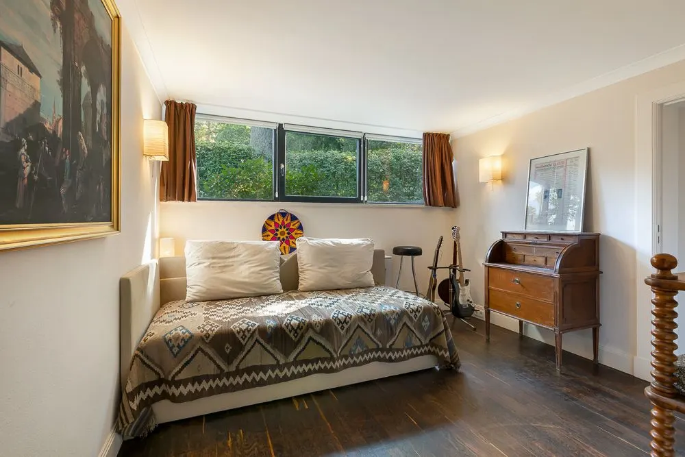 Zimmer mit Tagesbett, großem Fenster, gemusterter Decke, Schreibtisch und Musikinstrumenten.