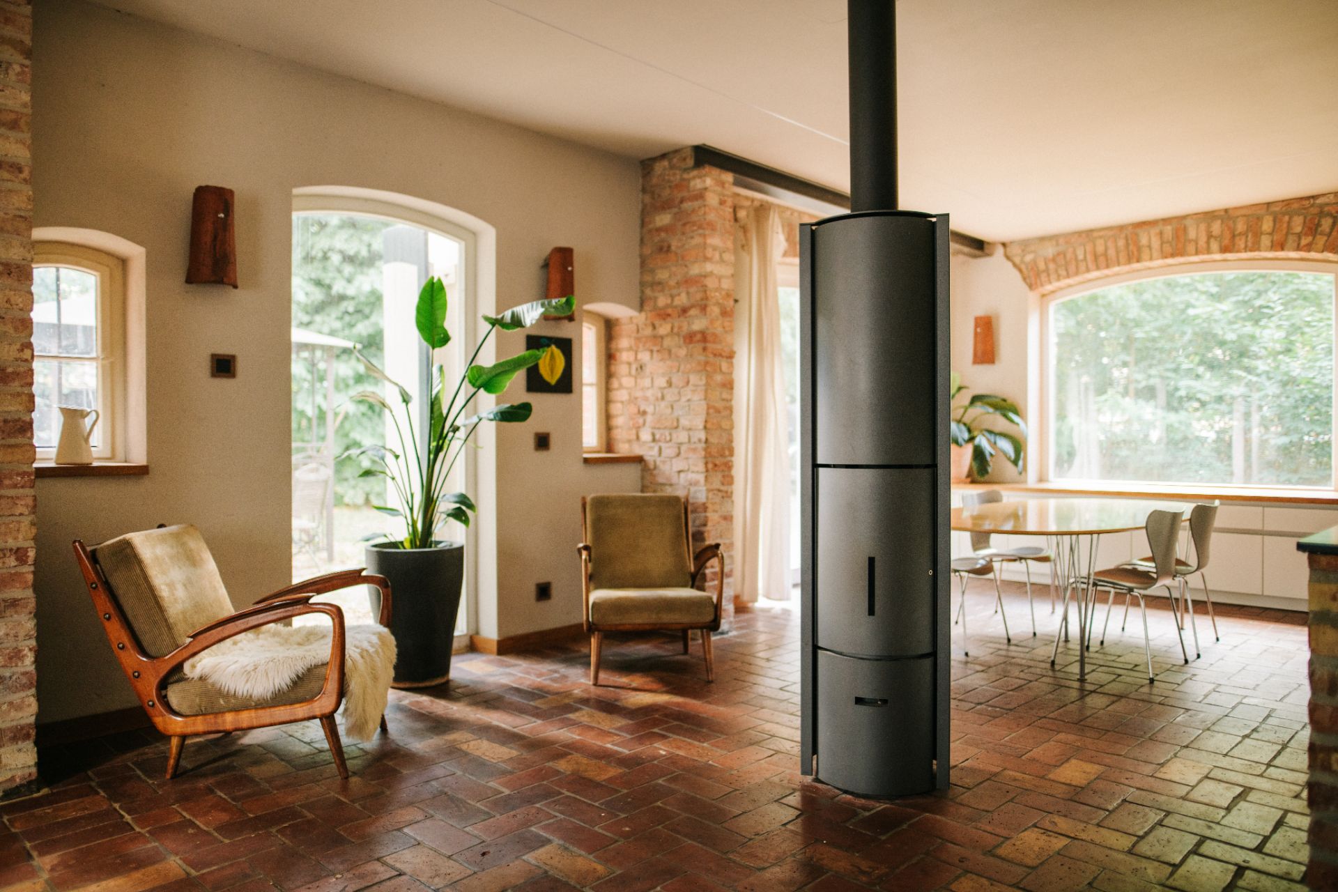 Wohnzimmer mit zwei Sesseln, einem Ofen in der Mitte und Essbereich mit Tisch und Stühlen am Fenster.