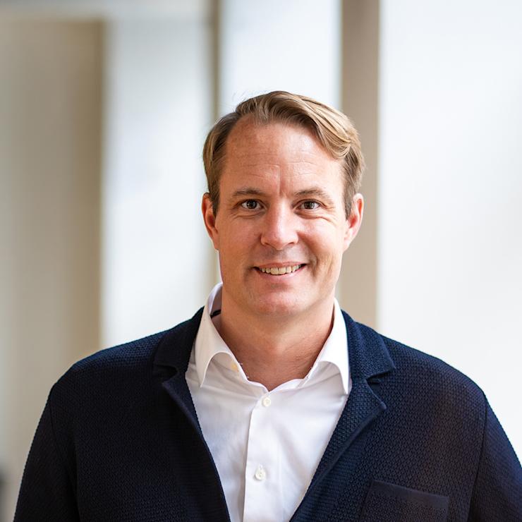 Mann in dunkler Jacke und weißem Hemd steht vor hellen Fenstern.