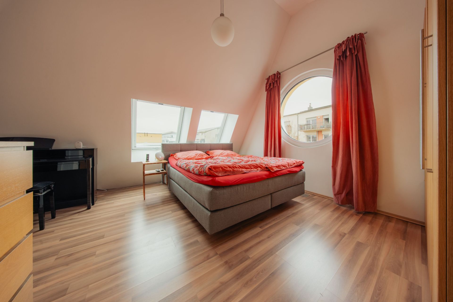 Schlafzimmer mit Doppelbett, rundem Fenster, roten Vorhängen und Holzboden.