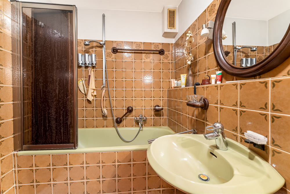 Kleines Badezimmer mit braunen Fliesen, Badewanne, Waschbecken und rundem Spiegel.