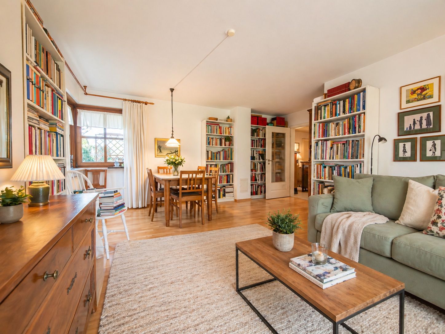 Helles Wohnzimmer mit Sofa, Couchtisch, Bücherregalen, Esstisch und großen Fenstern.
