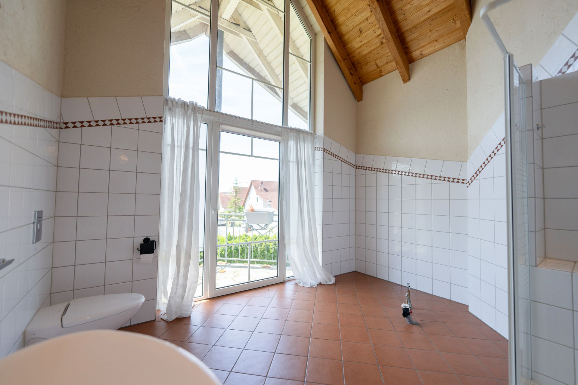 Helles Bad mit Fliesenboden, großer Fensterfront, WC und Duschbereich unter Holzdecke.