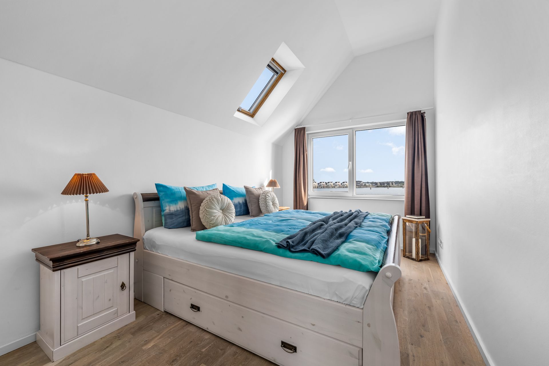 Helles Schlafzimmer mit Doppelbett, Nachttischlampe, Holzfußboden, Dachfenster und großem Fenster.