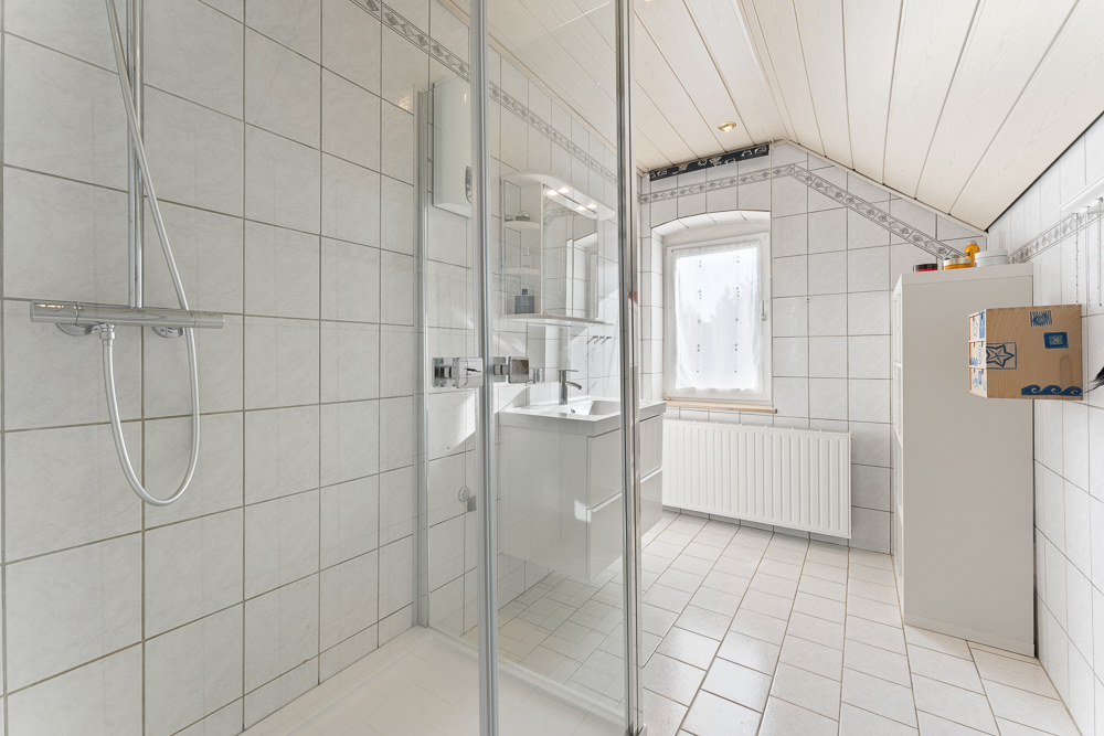 Helles Badezimmer mit Dusche, Waschbecken, Fenster und weiß gefliestem Boden.