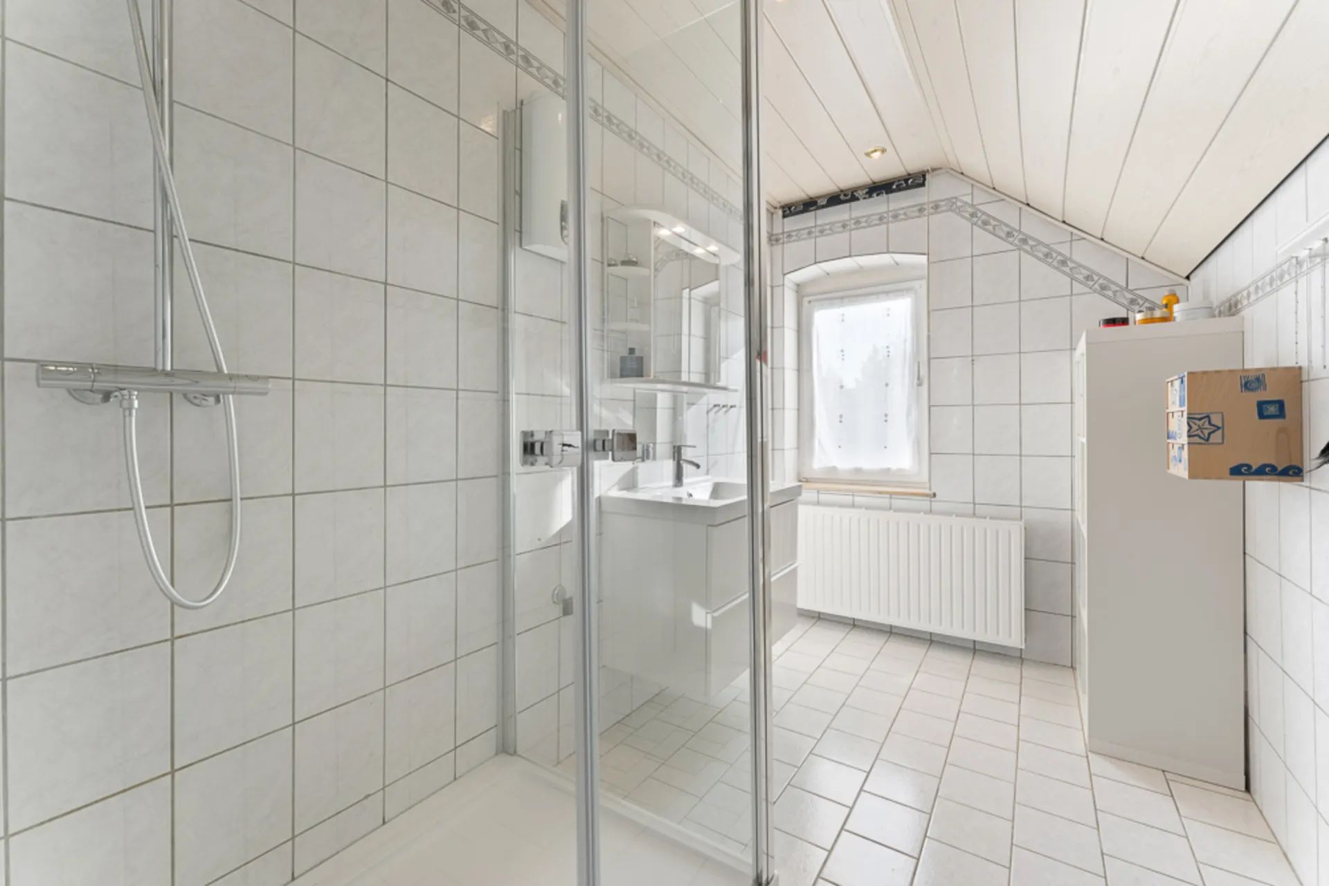 Helles Badezimmer mit Dusche, Waschbecken, Fenster und weiß gefliestem Boden.