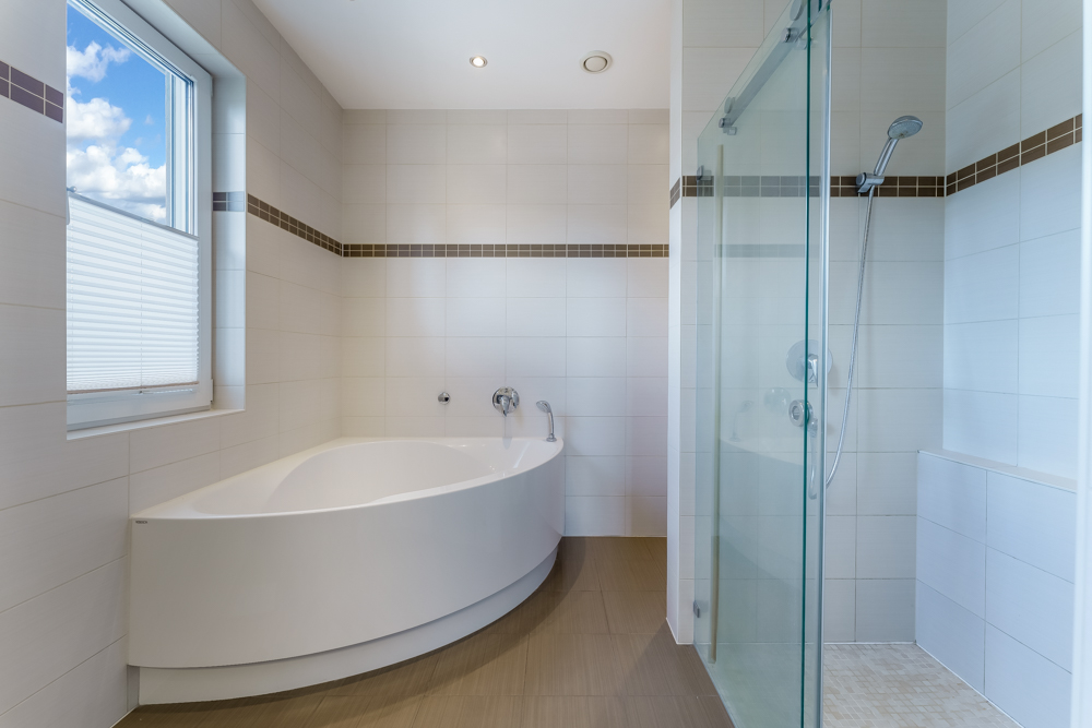 Helles Badezimmer mit Badewanne, Dusche mit Glaswand und Fenster mit Jalousie.