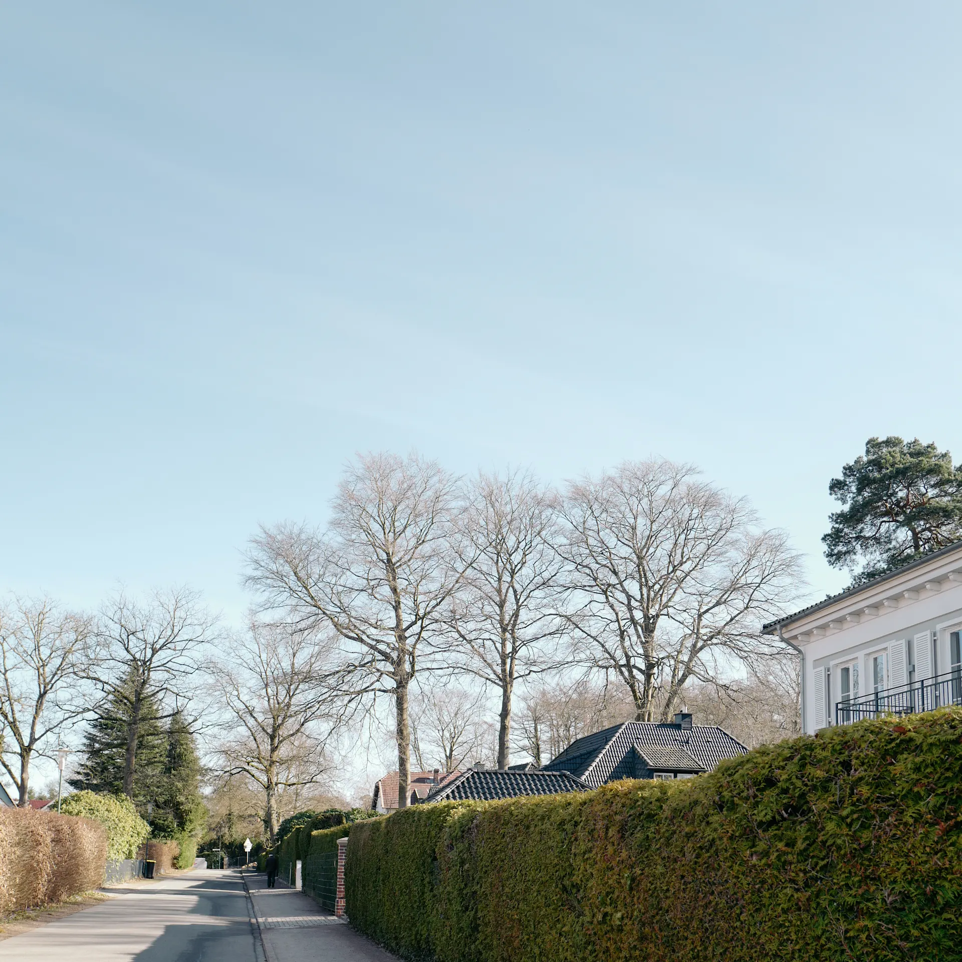 Ruhige Wohnstraße mit Bürgersteig, Hecken und Haus im Hintergrund bei klarem Himmel.