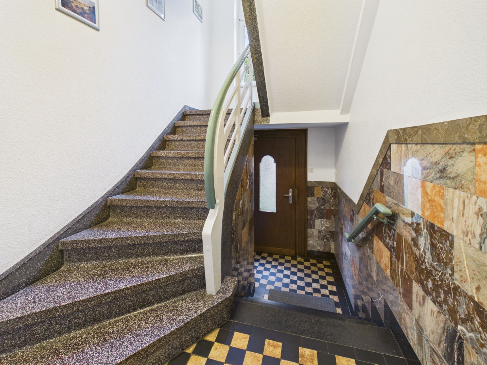 Treppenhaus mit Steinfliesen, Handlauf und Holztür im Eingangsbereich eines Wohnhauses.