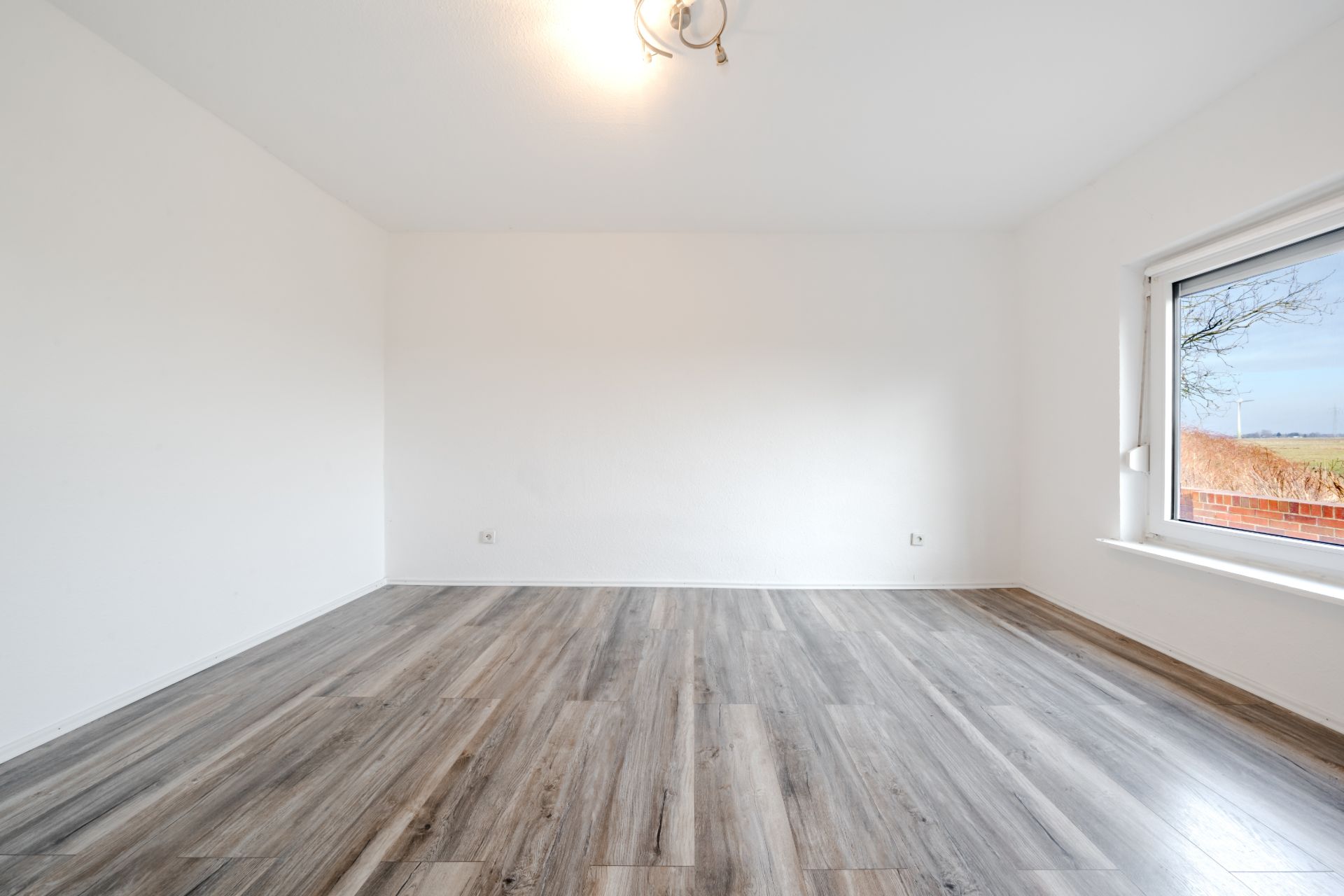 Leerer Raum mit hellem Holzboden, weißen Wänden und großem Fenster.