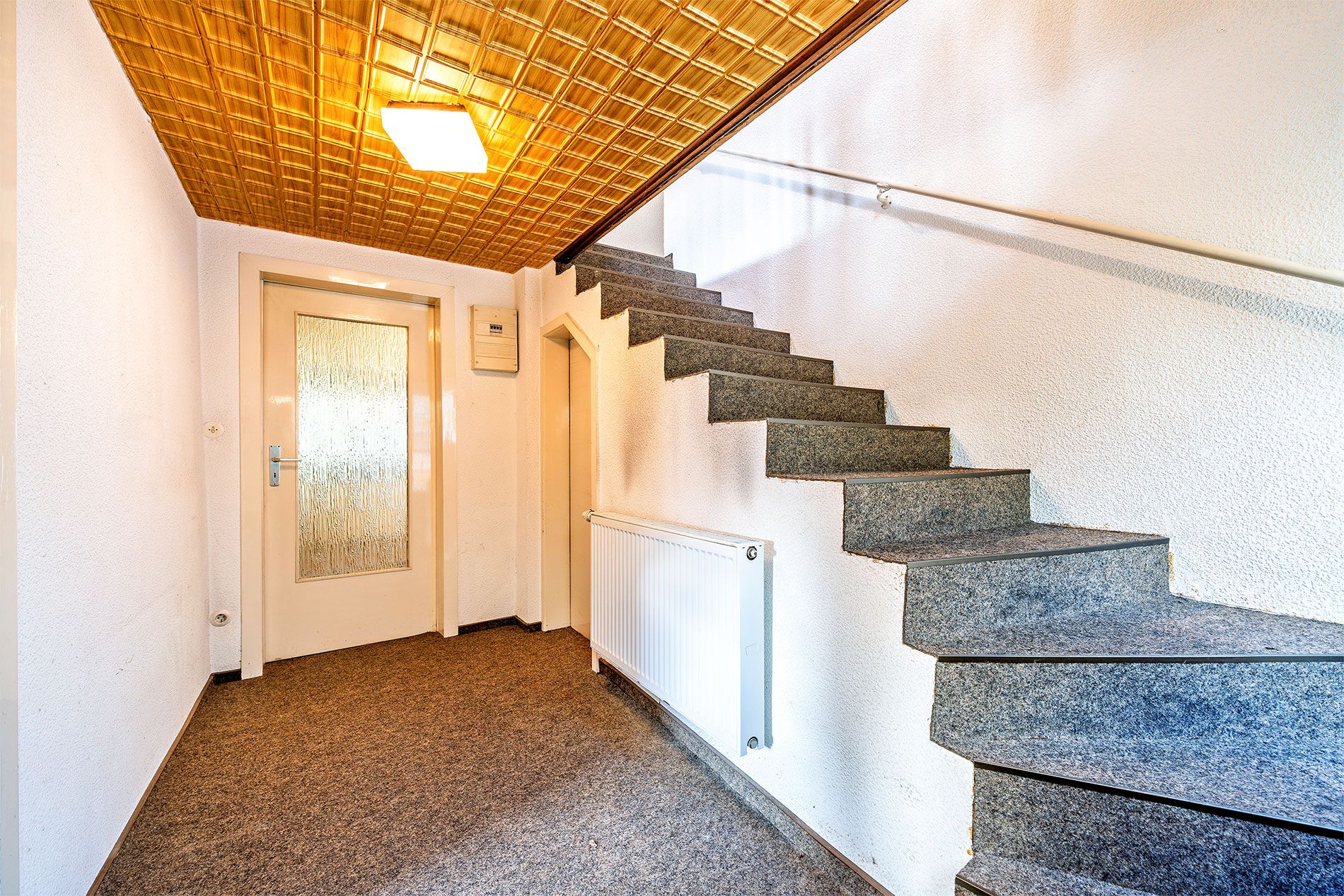 Flur mit Teppichboden, Treppe aus Stein, Holzbalkondecke und weißer Tür mit Glaseinsatz.