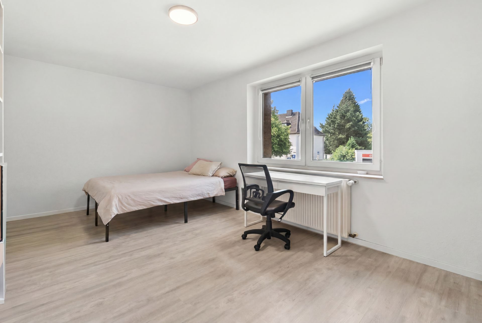 Helles Schlafzimmer mit Bett, Schreibtisch und Bürostuhl vor großem Fenster.