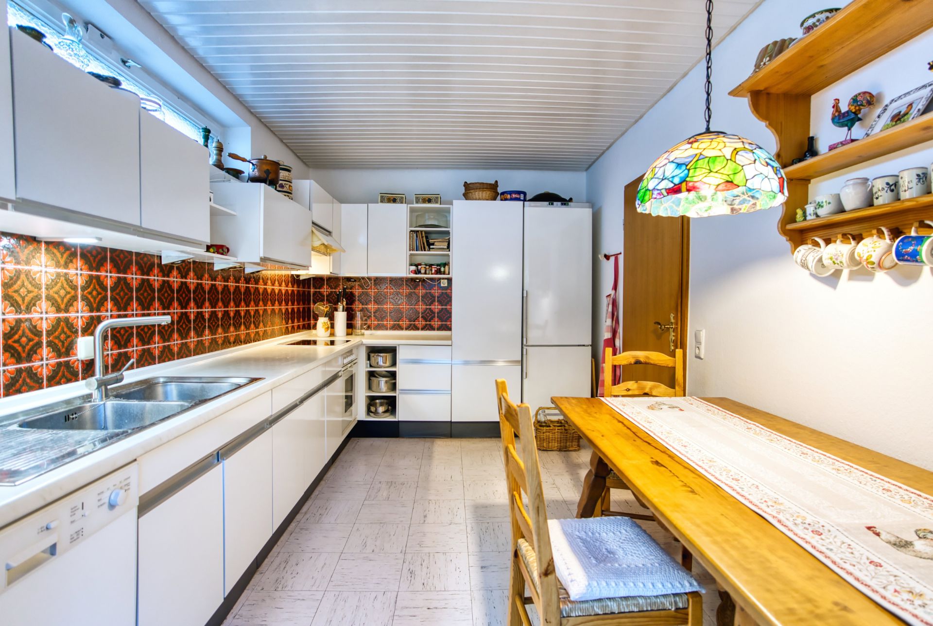 Küche mit weißen Schränken, Holz-Esstisch, Spüle, bunter Lampe und Wandregalen mit Geschirr.