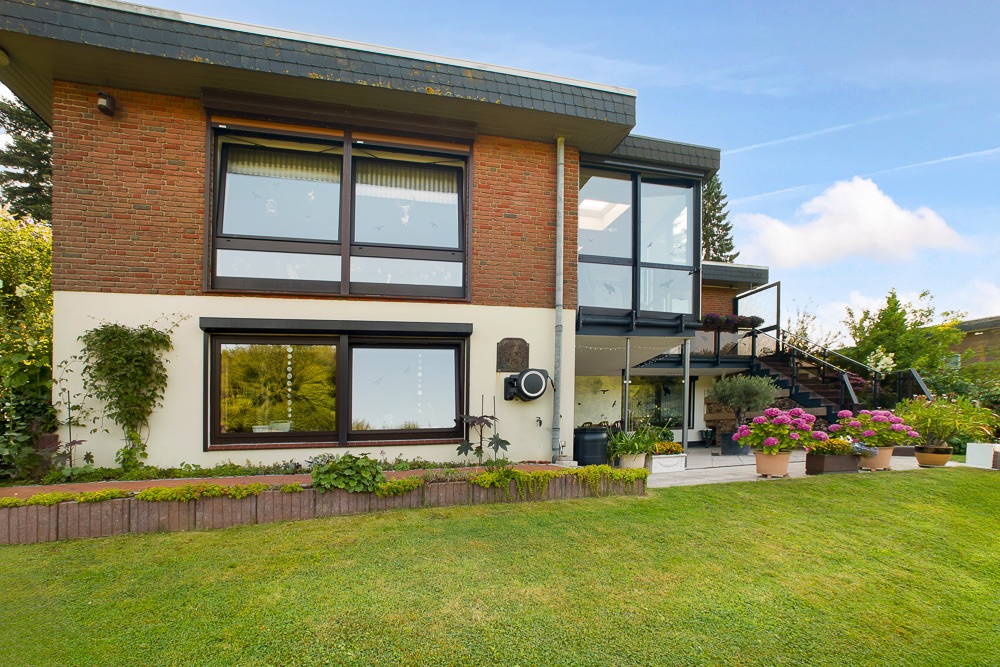 Modernes zweistöckiges Haus mit großen Fenstern, Terrasse, Garten und Blumenbeeten.