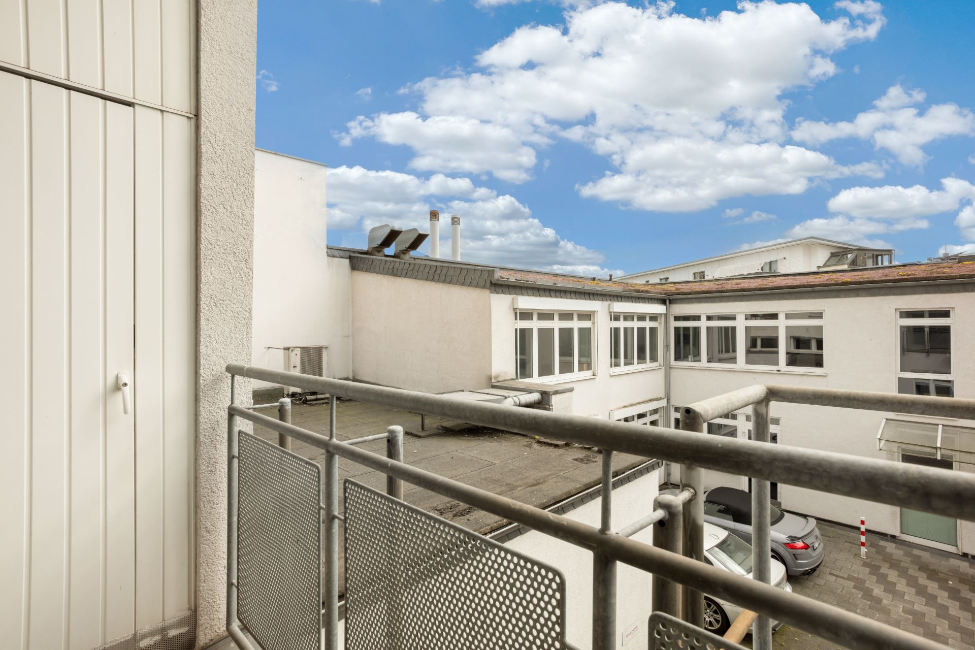 Blick von einem Balkon auf einen Innenhof mit geparktem Auto und weißen Gebäuden.