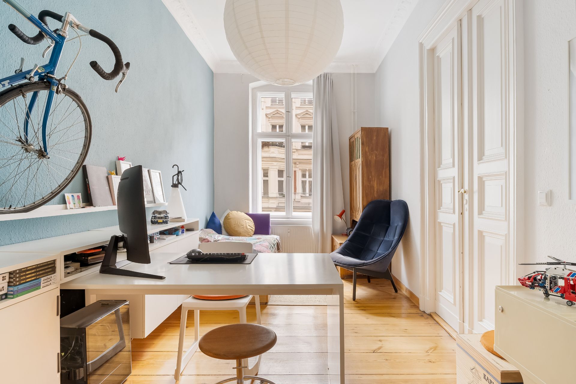 Helles Arbeitszimmer mit Schreibtisch, Computer, Stuhl, Regal, Wandfahrrad und großem Fenster.