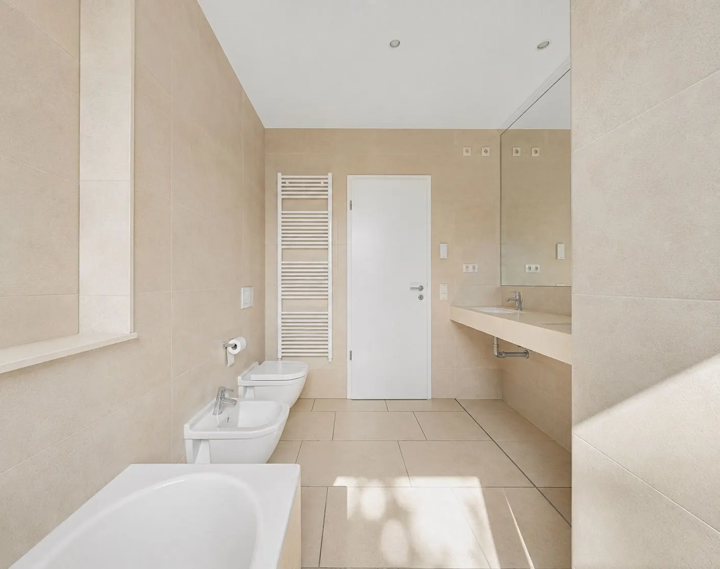Modernes, helles Badezimmer mit Badewanne, Bidet, WC, großem Spiegel und Handtuchheizkörper.