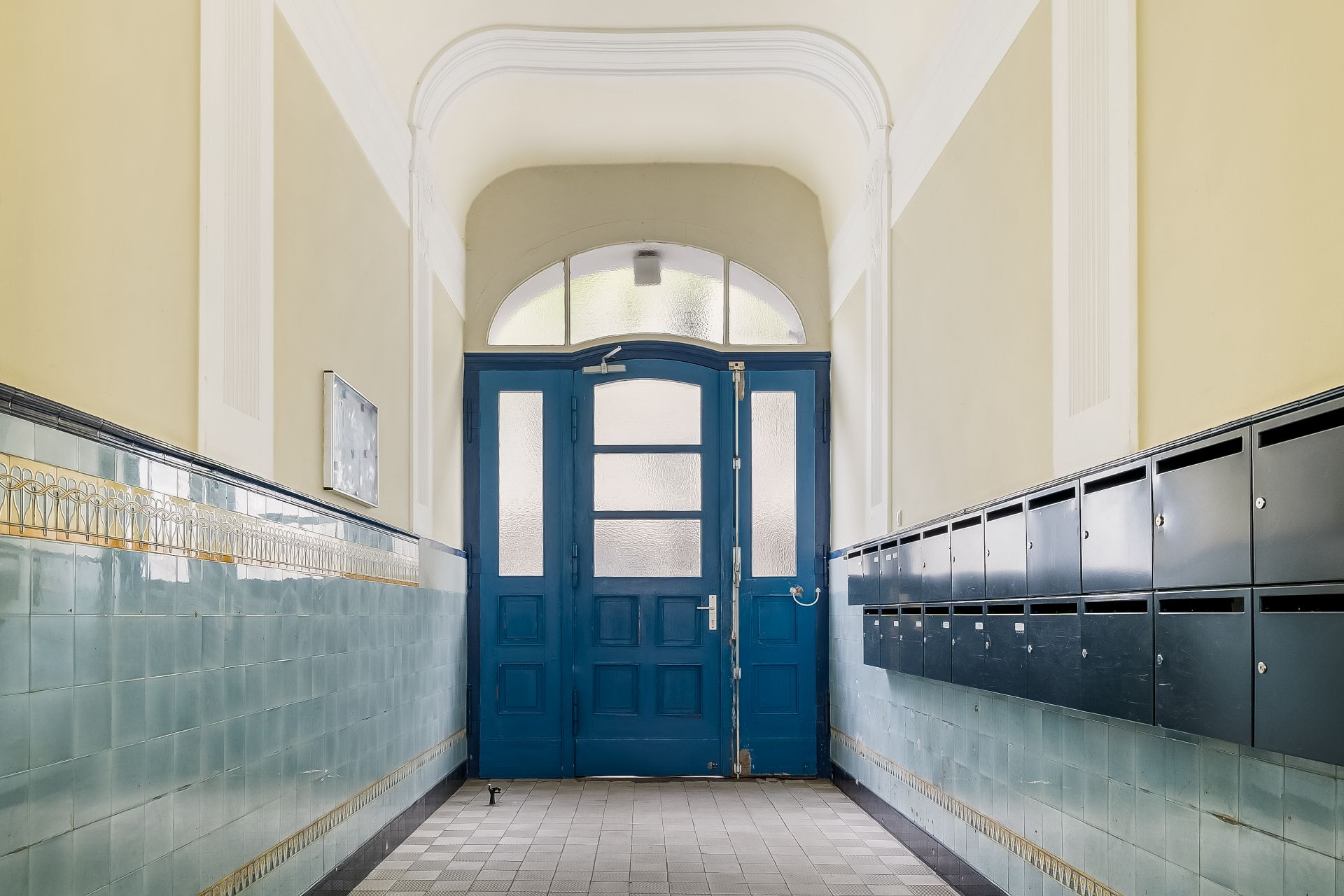 Hausflur mit blauer Eingangstür, Fliesenwänden und Briefkästen auf der rechten Seite.