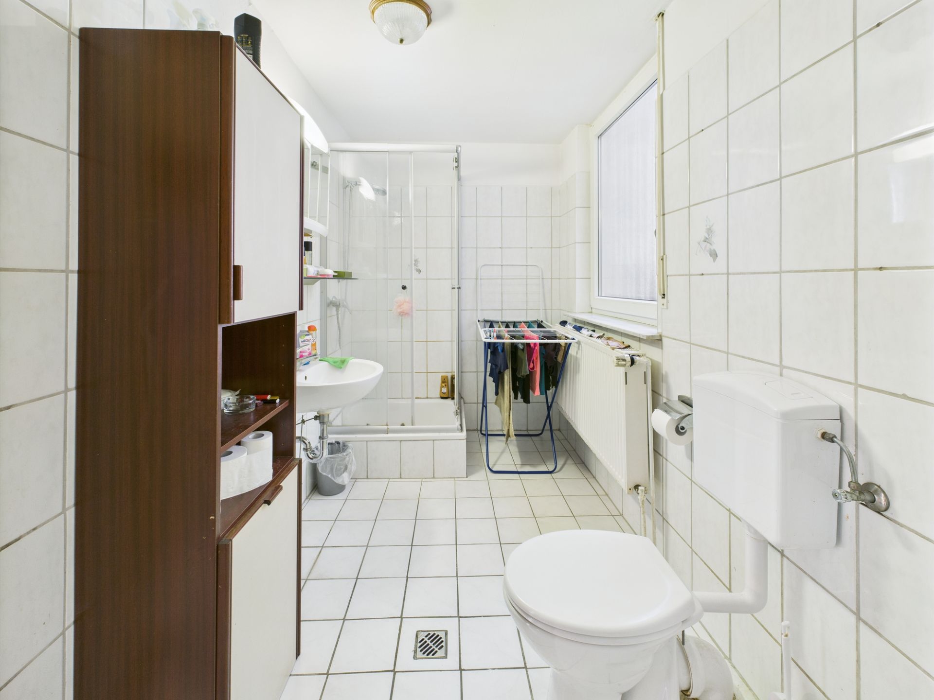 Helles Badezimmer mit Dusche, WC, Waschbecken, Schrank und Wäscheständer vor dem Fenster.