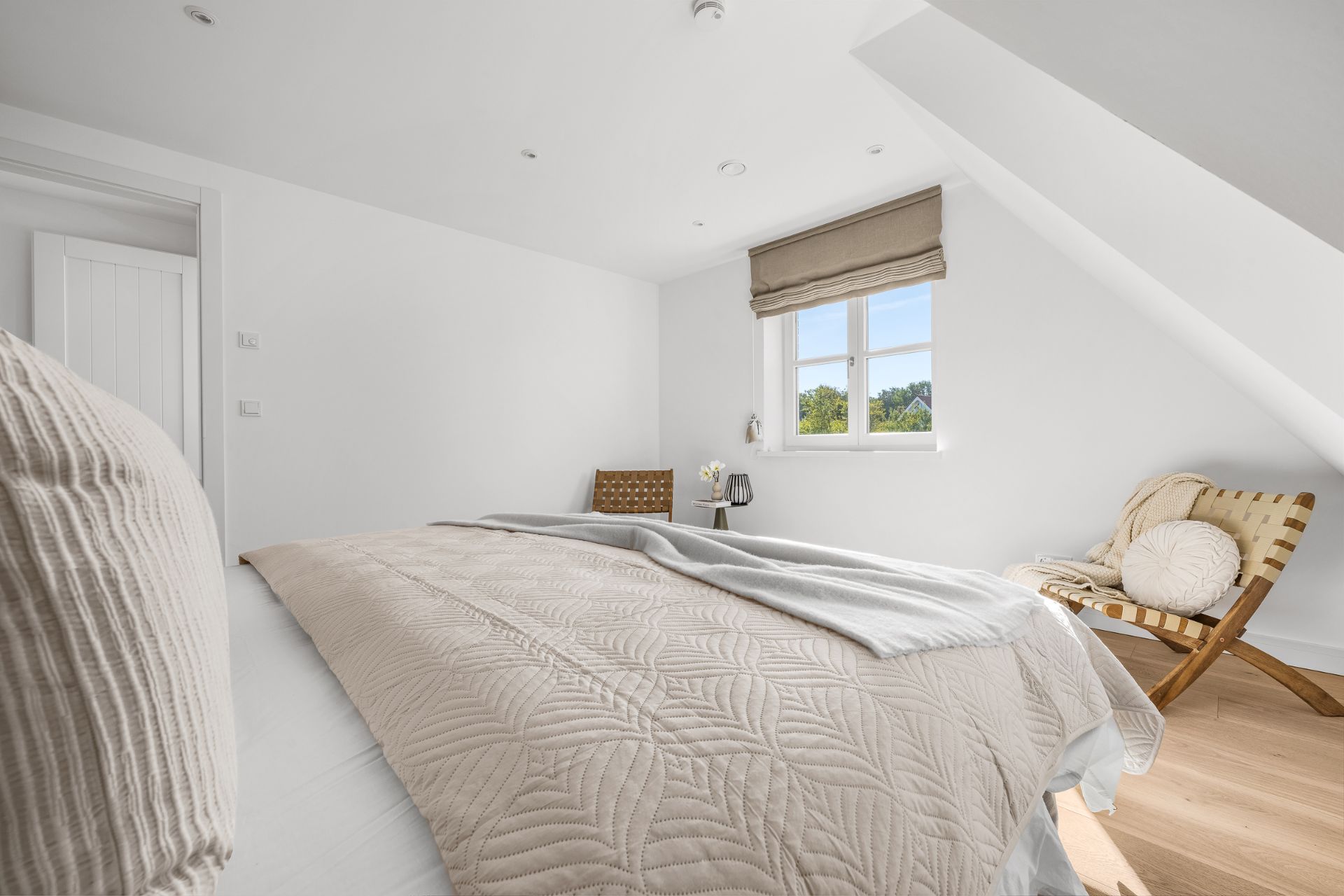 Helles Schlafzimmer mit Doppelbett, Holzstuhl, Fenster und hellem Holzboden.