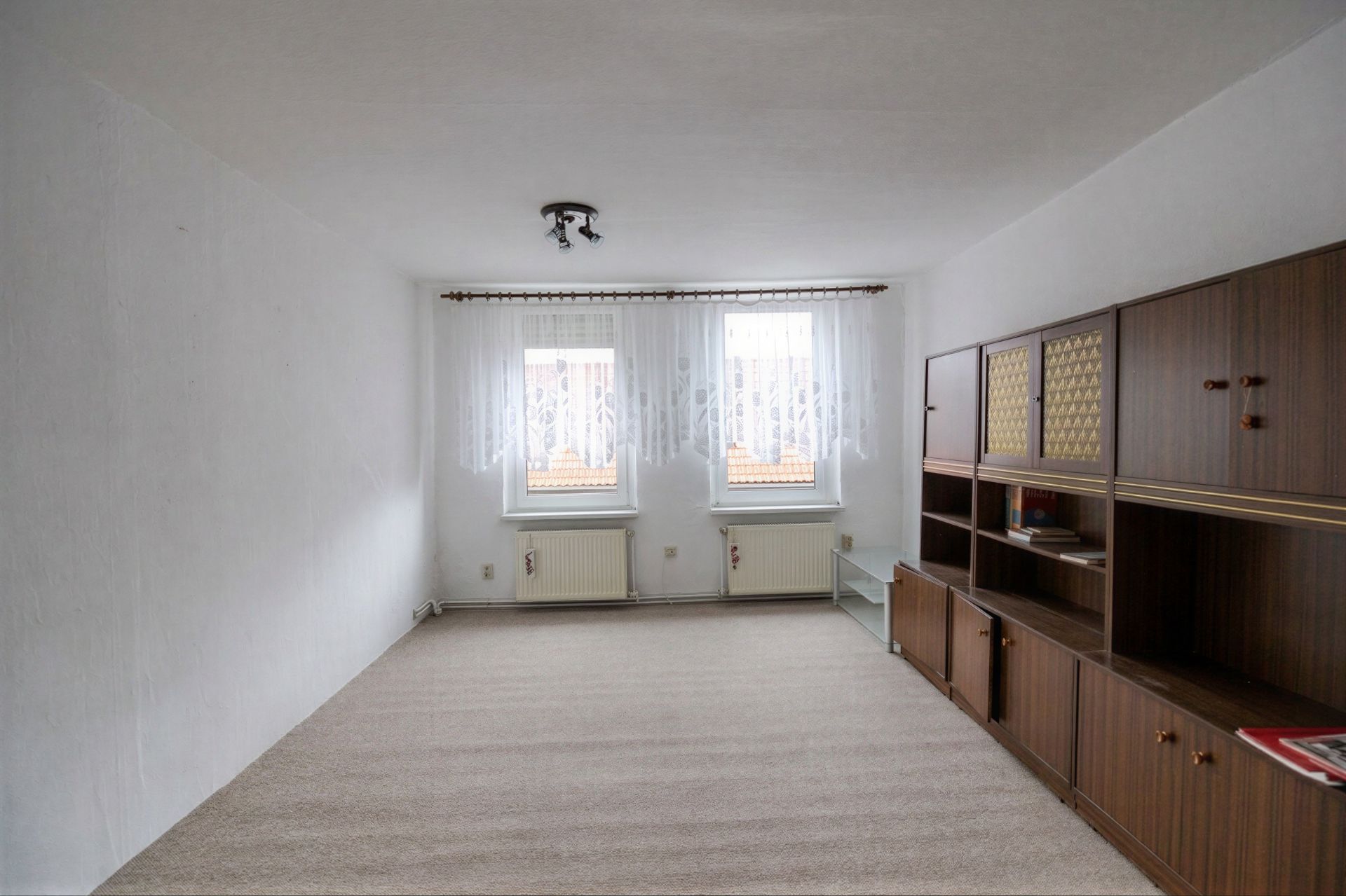 Helles Wohnzimmer mit Teppichboden, zwei Fenstern und großer Holzschrankwand.