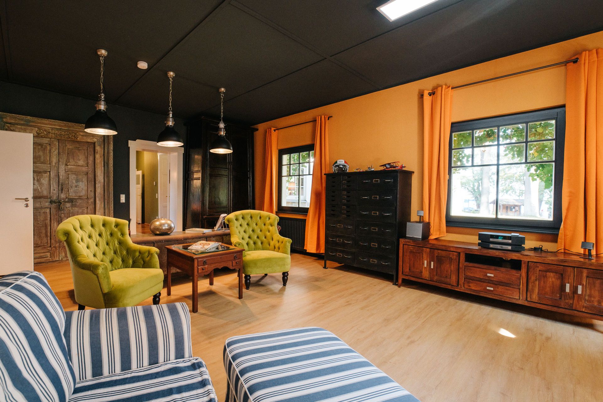 Wohnzimmer mit gelben Sesseln, gestreiftem Sofa, Holzmöbeln und orangefarbenen Vorhängen.