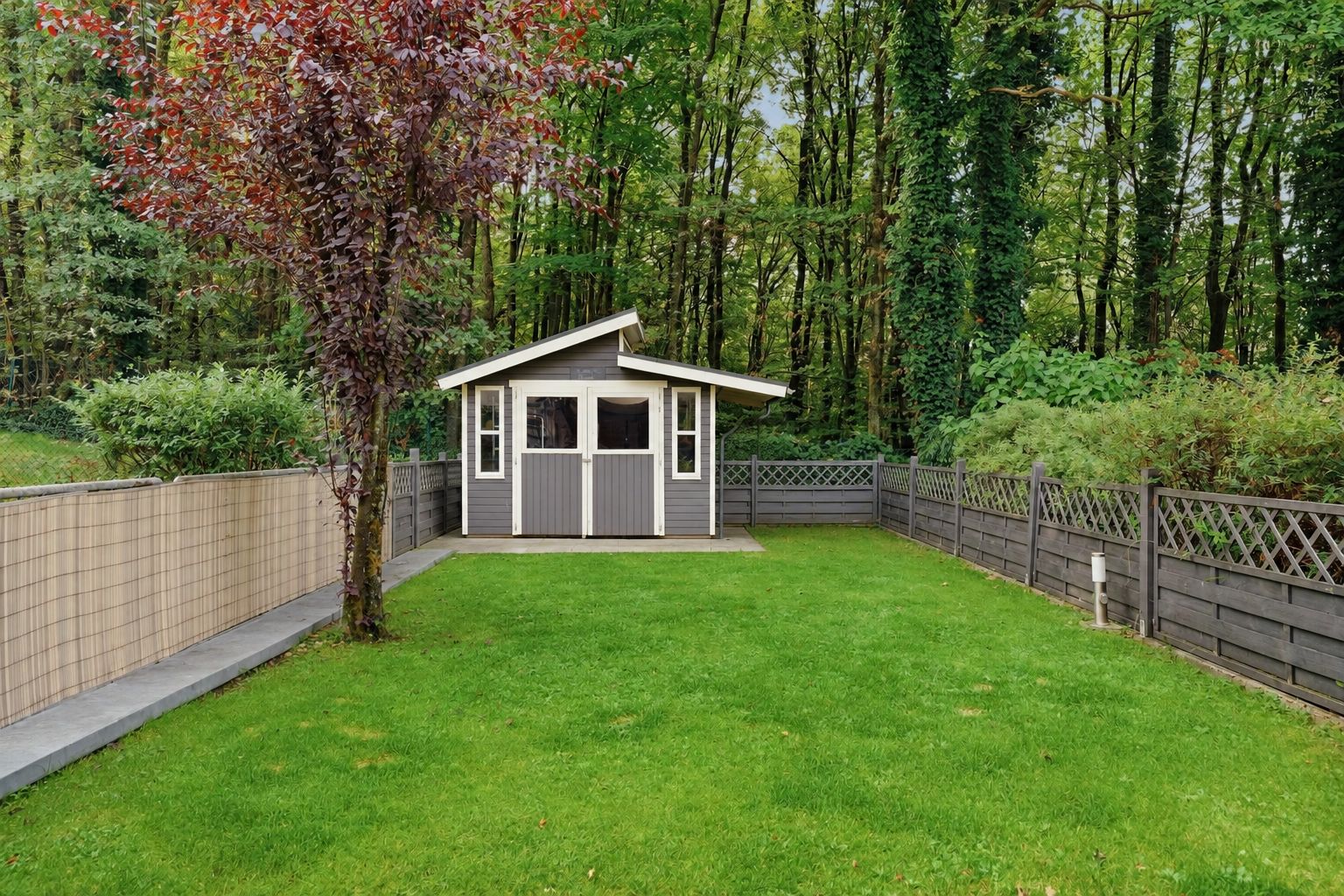 Holzgartenhaus auf grünem Rasen, umgeben von Zaun und Bäumen.