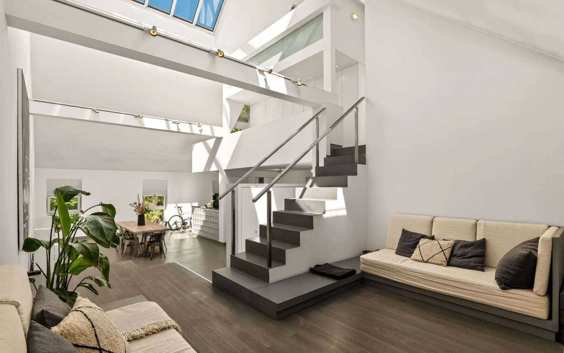 Heller Wohnraum mit zwei Sofas, Treppe, Esstisch und großen Fenstern.