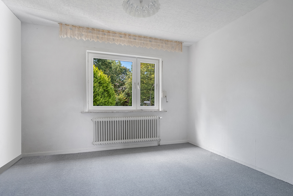 Leeres Zimmer mit Teppichboden, großem Fenster, Heizkörper und Blick ins Grüne.