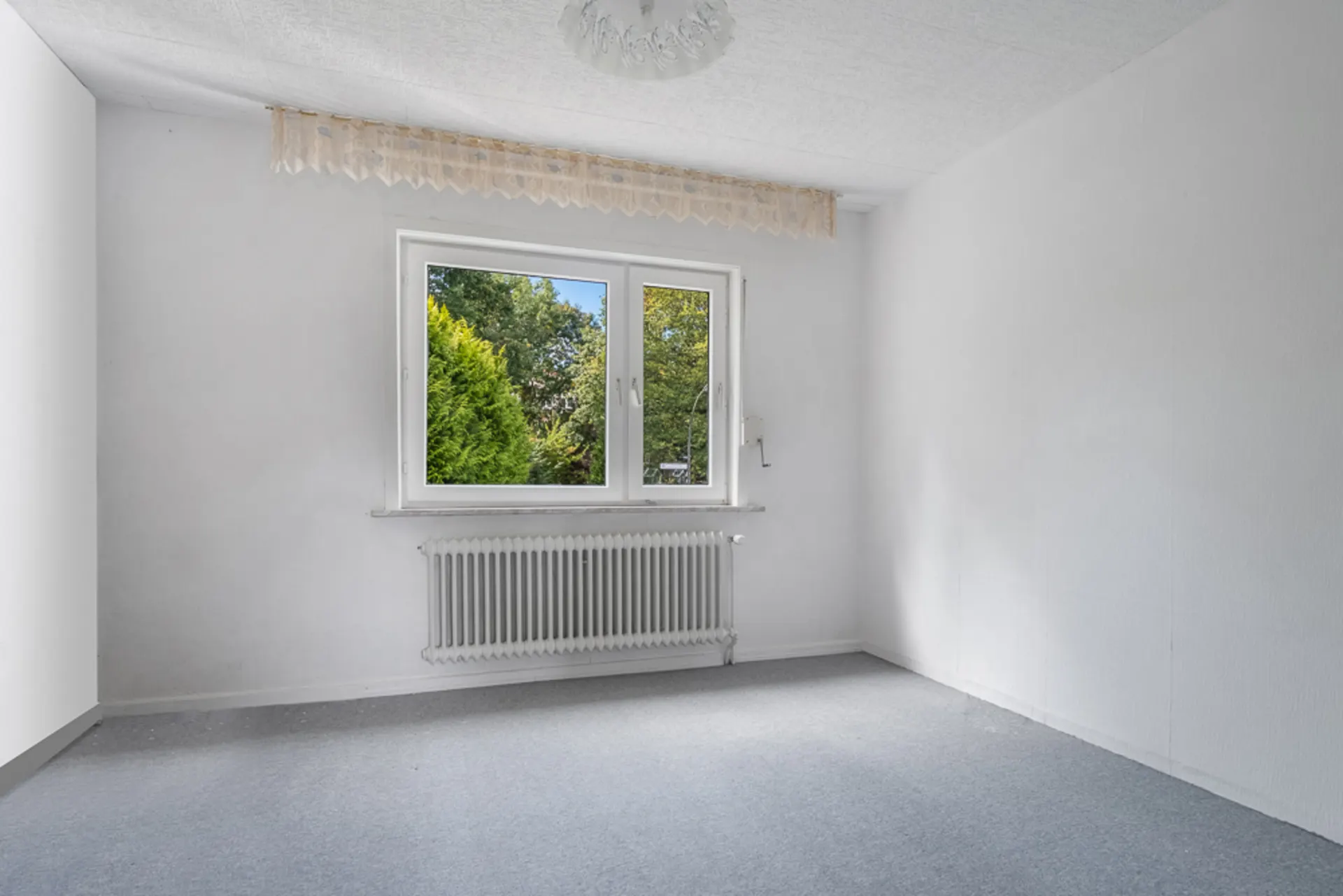 Leeres Zimmer mit Teppichboden, großem Fenster, Heizkörper und Blick ins Grüne.