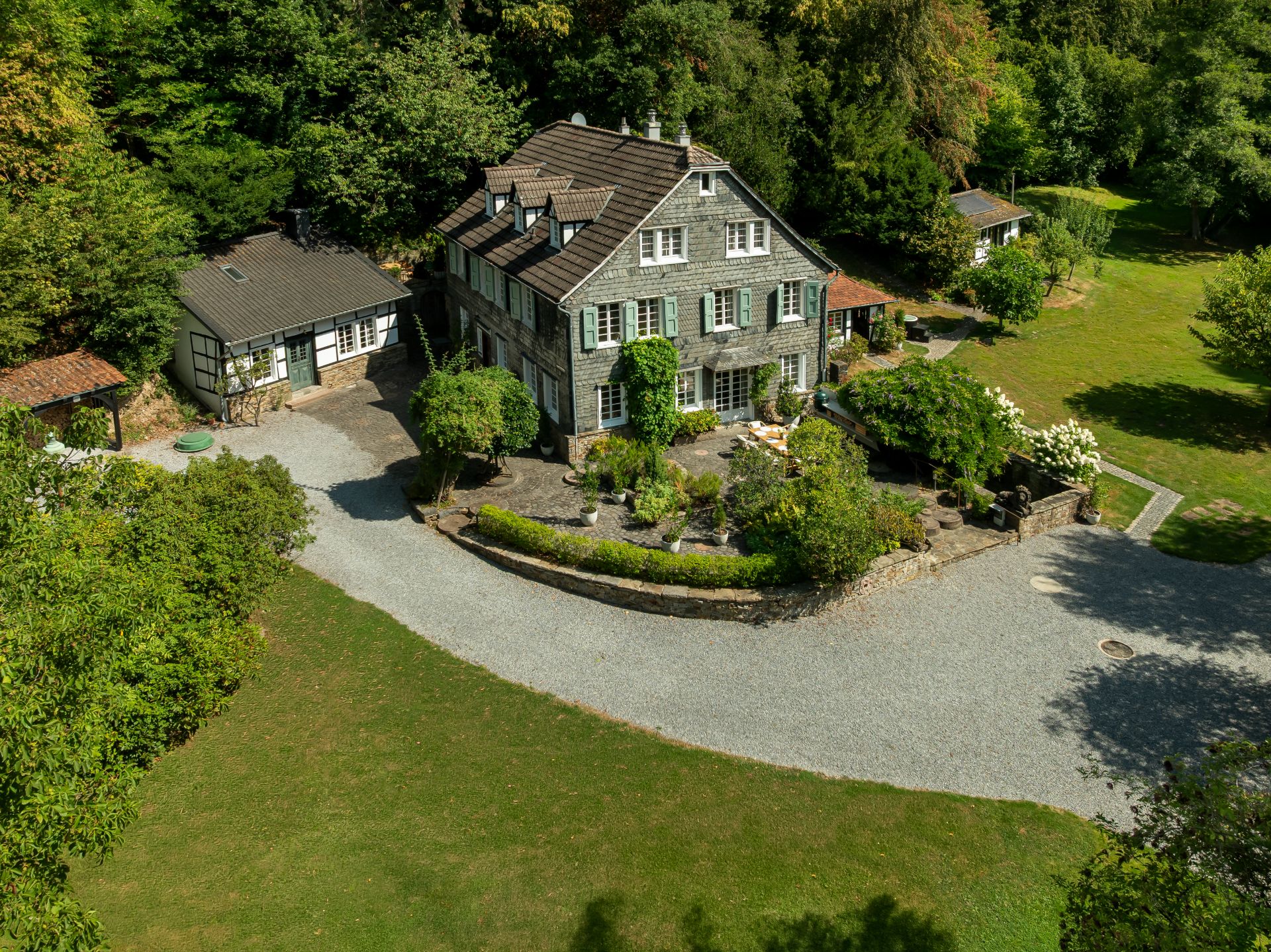 Großes Wohnhaus mit Garten, Nebengebäude und geschwungener Einfahrt, umgeben von Bäumen.