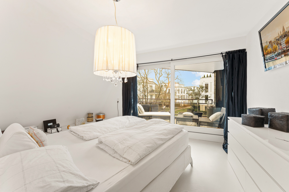 Helles Schlafzimmer mit Doppelbett, weißer Kommode und Zugang zu einem Balkon mit Sitzmöbeln.
