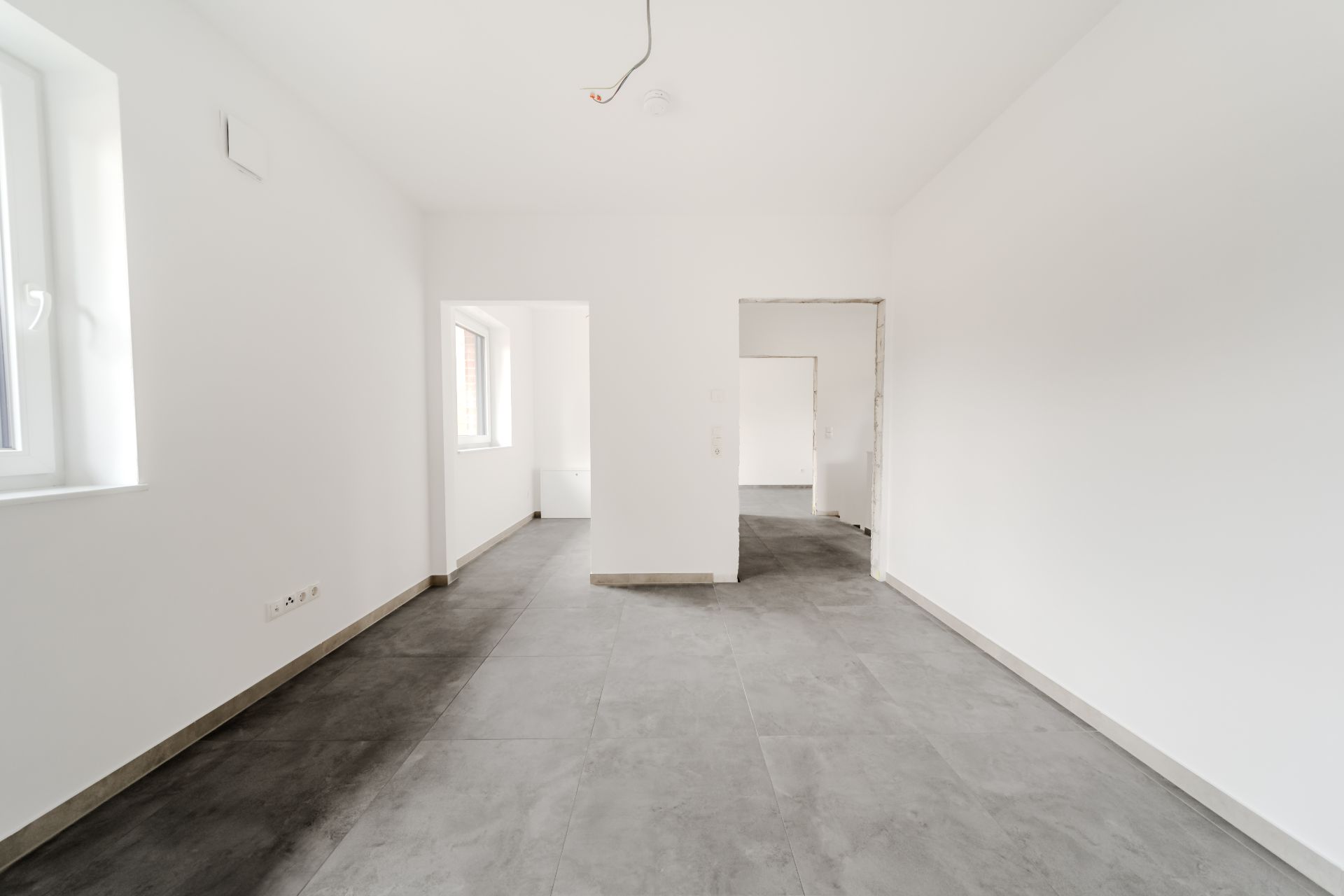 Leerer, heller Raum mit Fliesenboden und weißen Wänden, mehrere Türen und Fenster sichtbar.